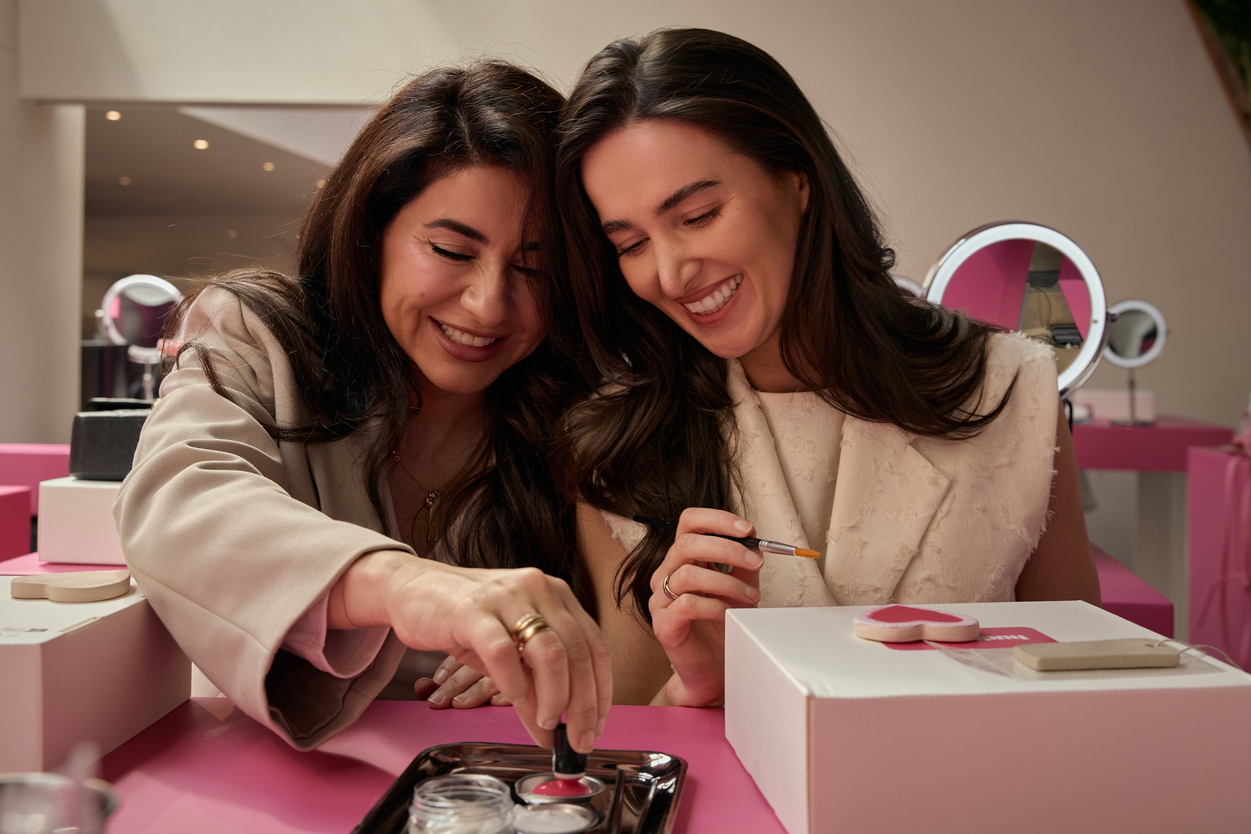 Two women happily painting cookies at a pink-themed table with makeup mirrors in the background.