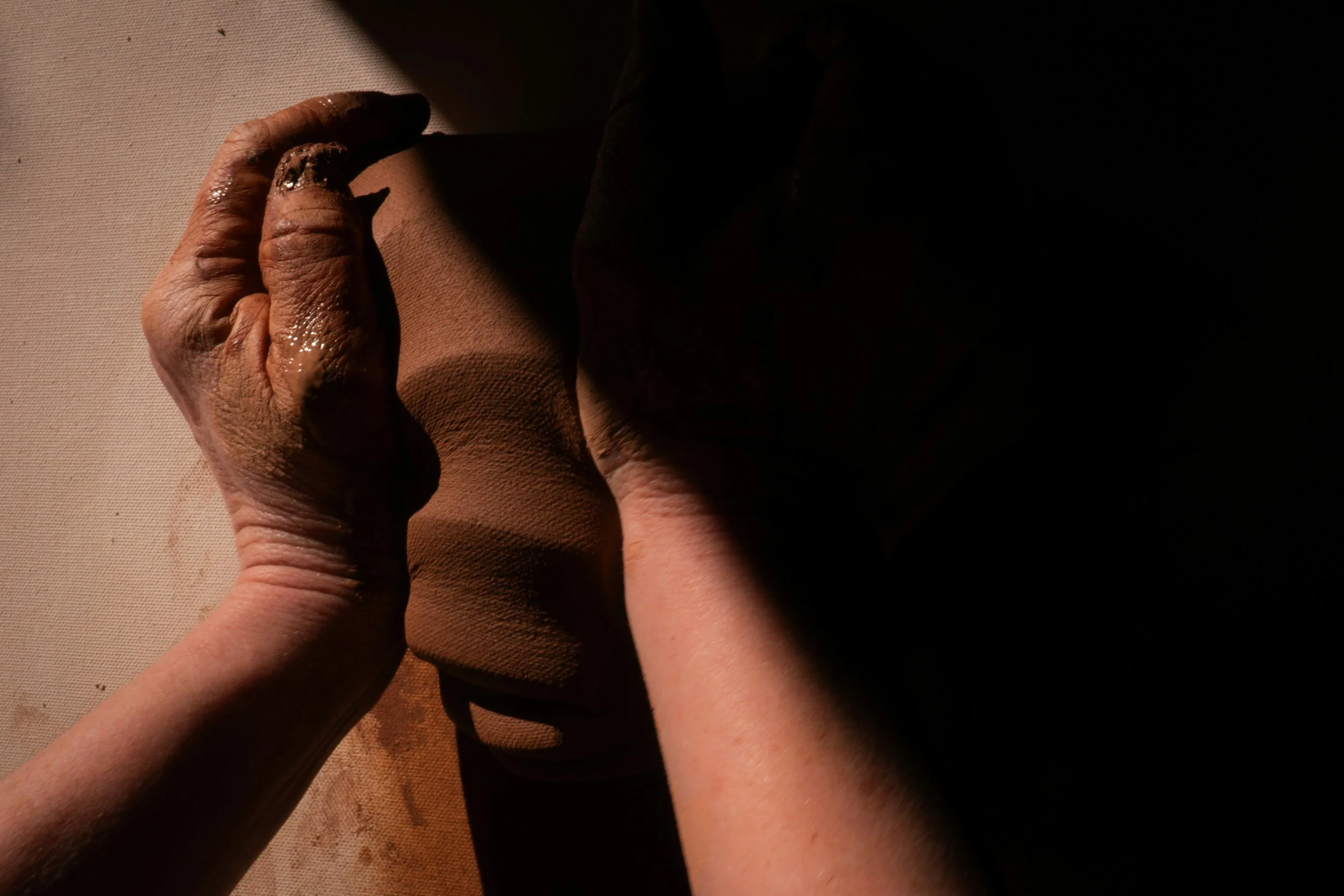 Close-up of two hands pressing clay on a surface, with shadows and light highlighting the texture of the hands and clay.