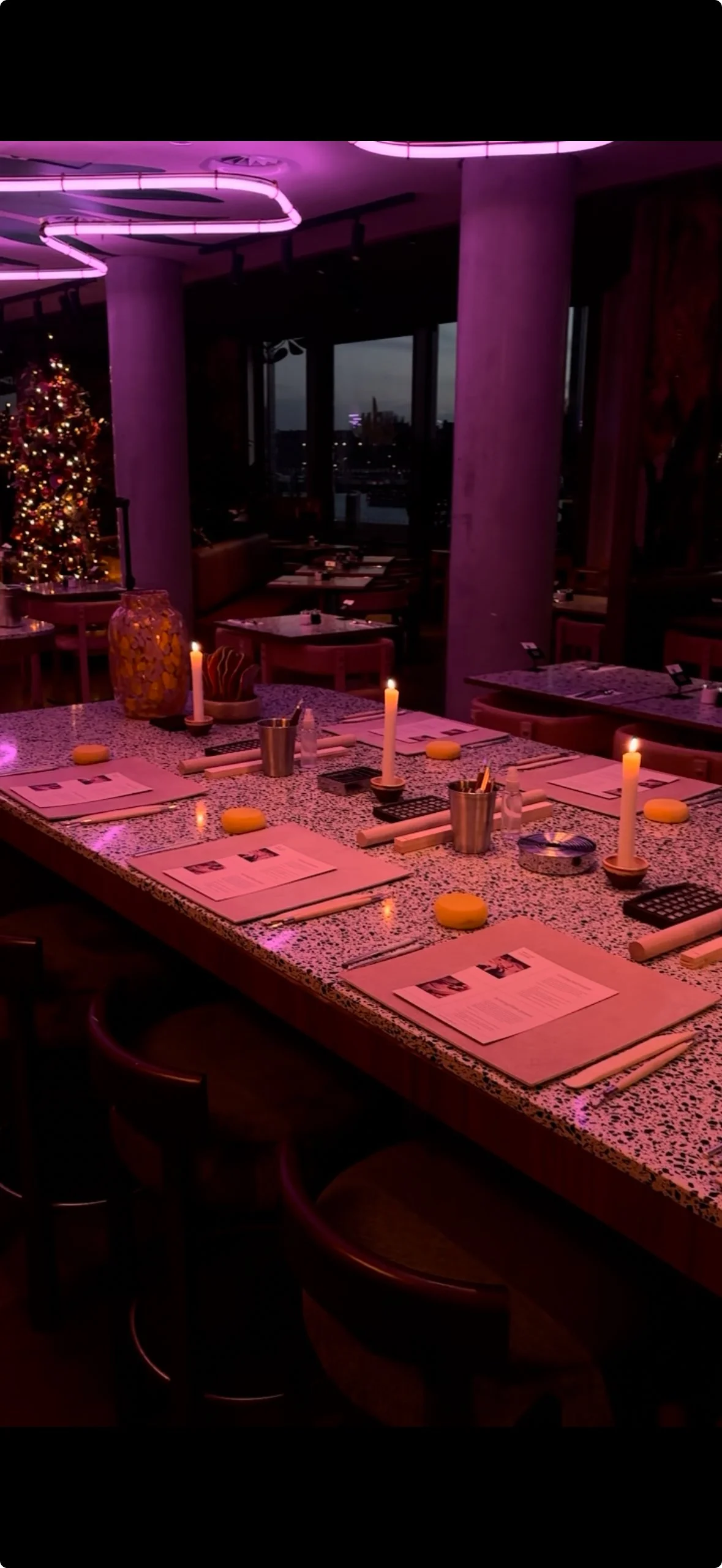 A restaurant with a long speckled table set for a meal, illuminated by candlelight and purple neon lights, with a decorated Christmas tree in the background and windows showing an outdoor city view.