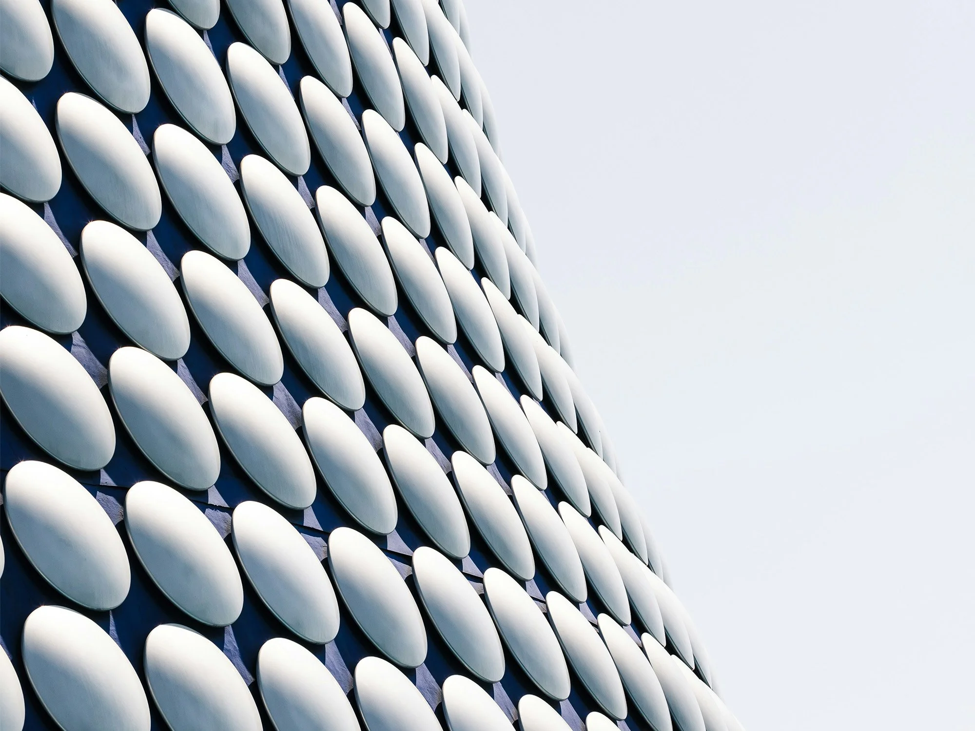 Close-up of a modern building's exterior with a pattern of silver, oval-shaped panels against a clear, light blue sky.