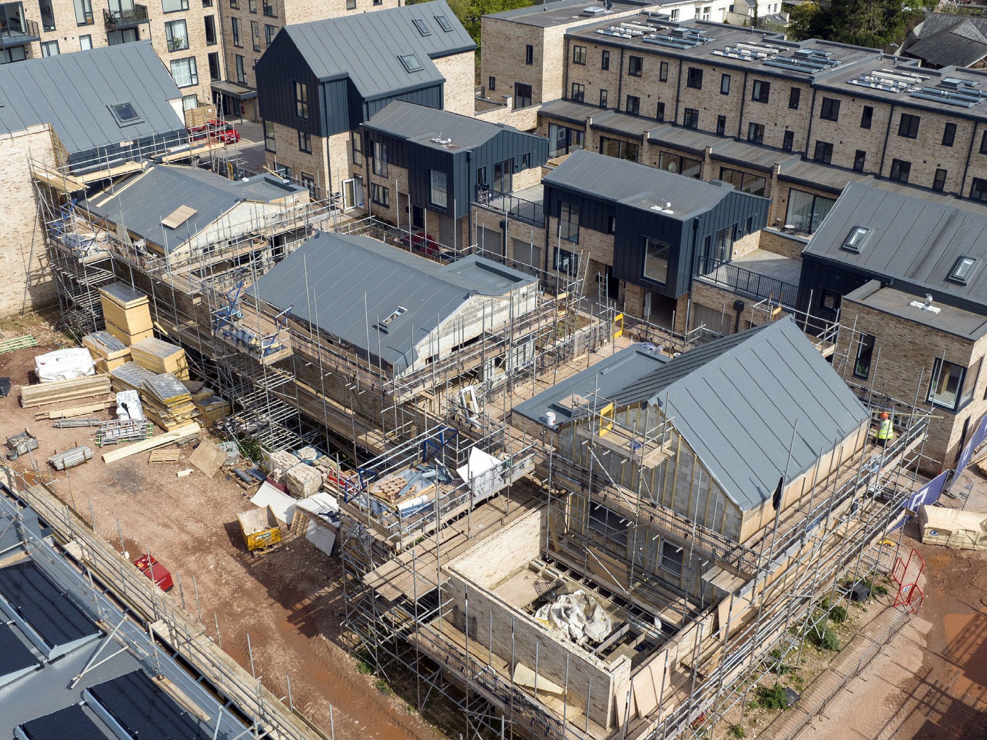 Aerial view of a construction site with partially built residential buildings surrounded by scaffolding and construction materials, with completed buildings in the background.