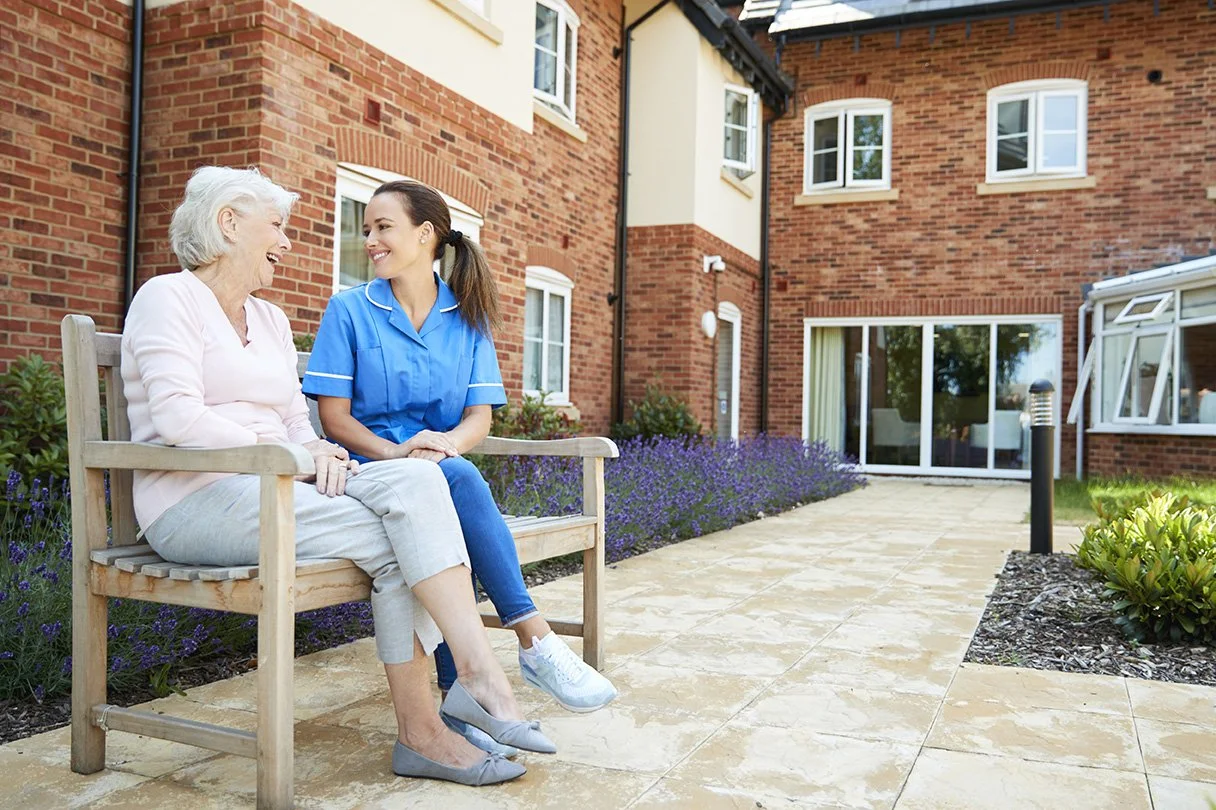 An elderly woman and a young female caregiver sitting on a bench outside a brick apartment building, smiling and engaging in conversation.