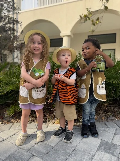 Three young children dressed as explorers with safari vests and hats, standing outdoors near greenery and a building, enjoying a pretend adventure.