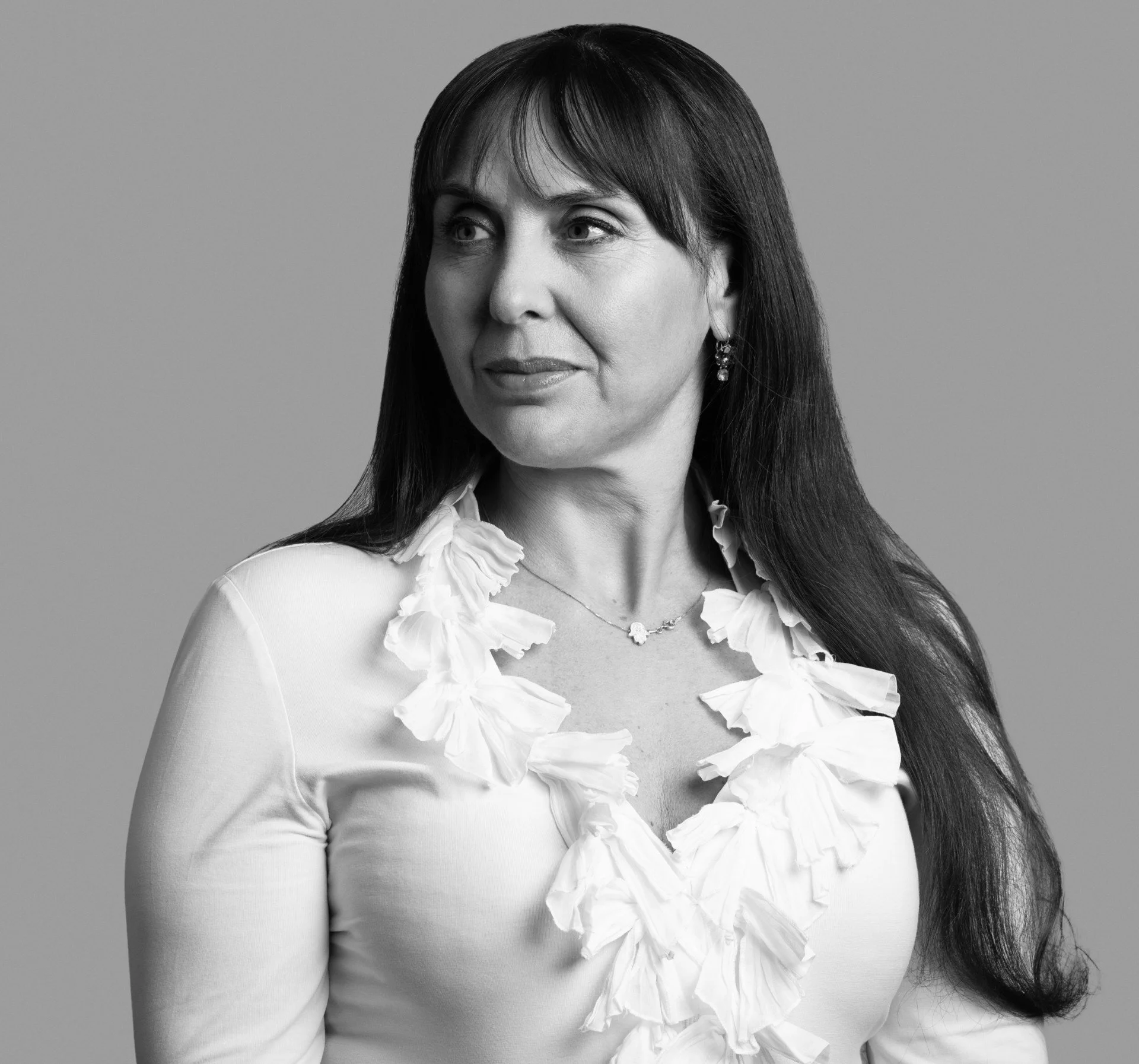 Black-and-white portrait of a woman with long dark hair, wearing earrings and a white top with ruffled details, against a plain background.