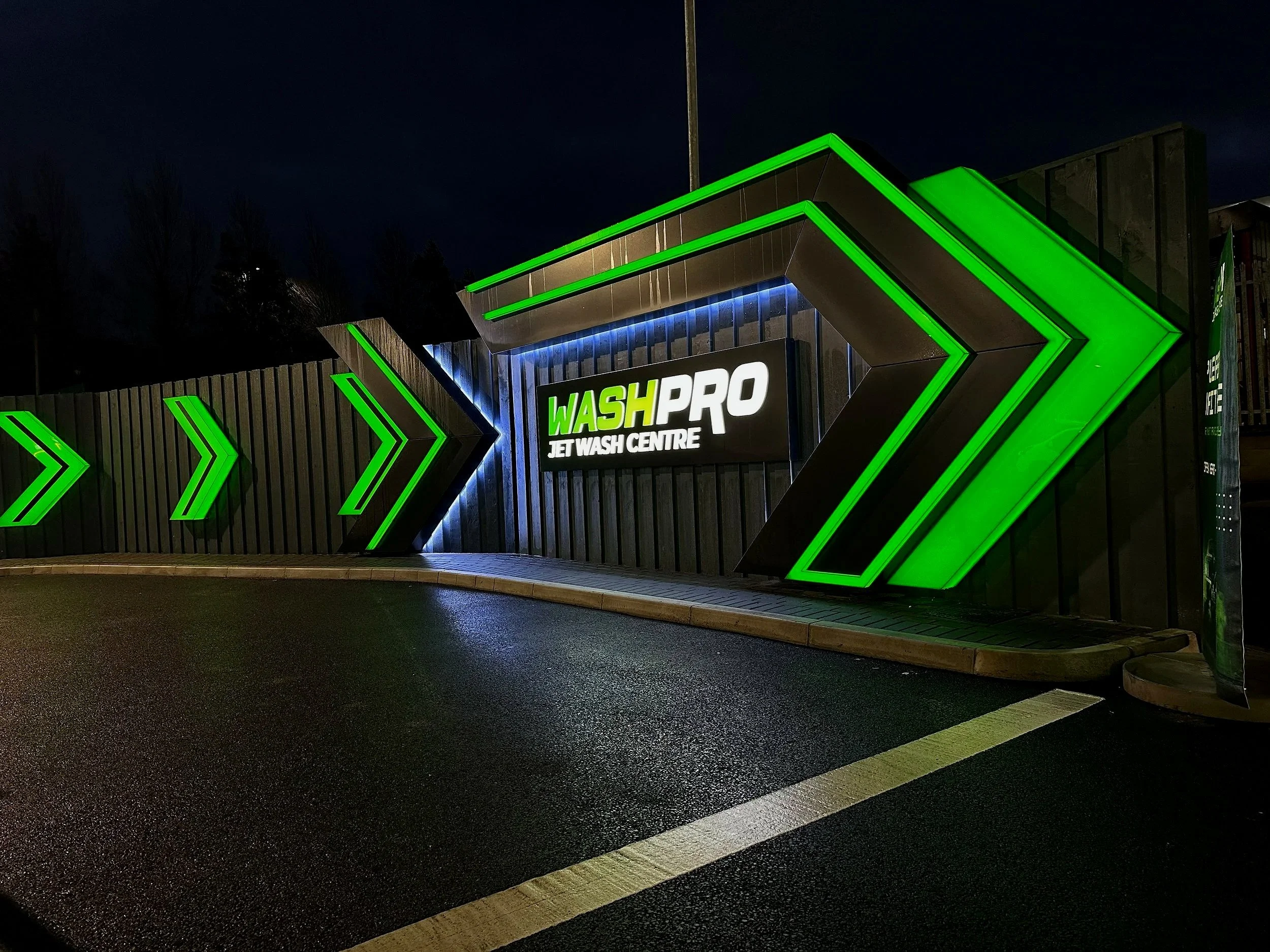 Night view of the WashPro Jet Wash Centre sign with illuminated green arrows pointing towards the entrance, black wall with vertical panels, and dark sky in the background.