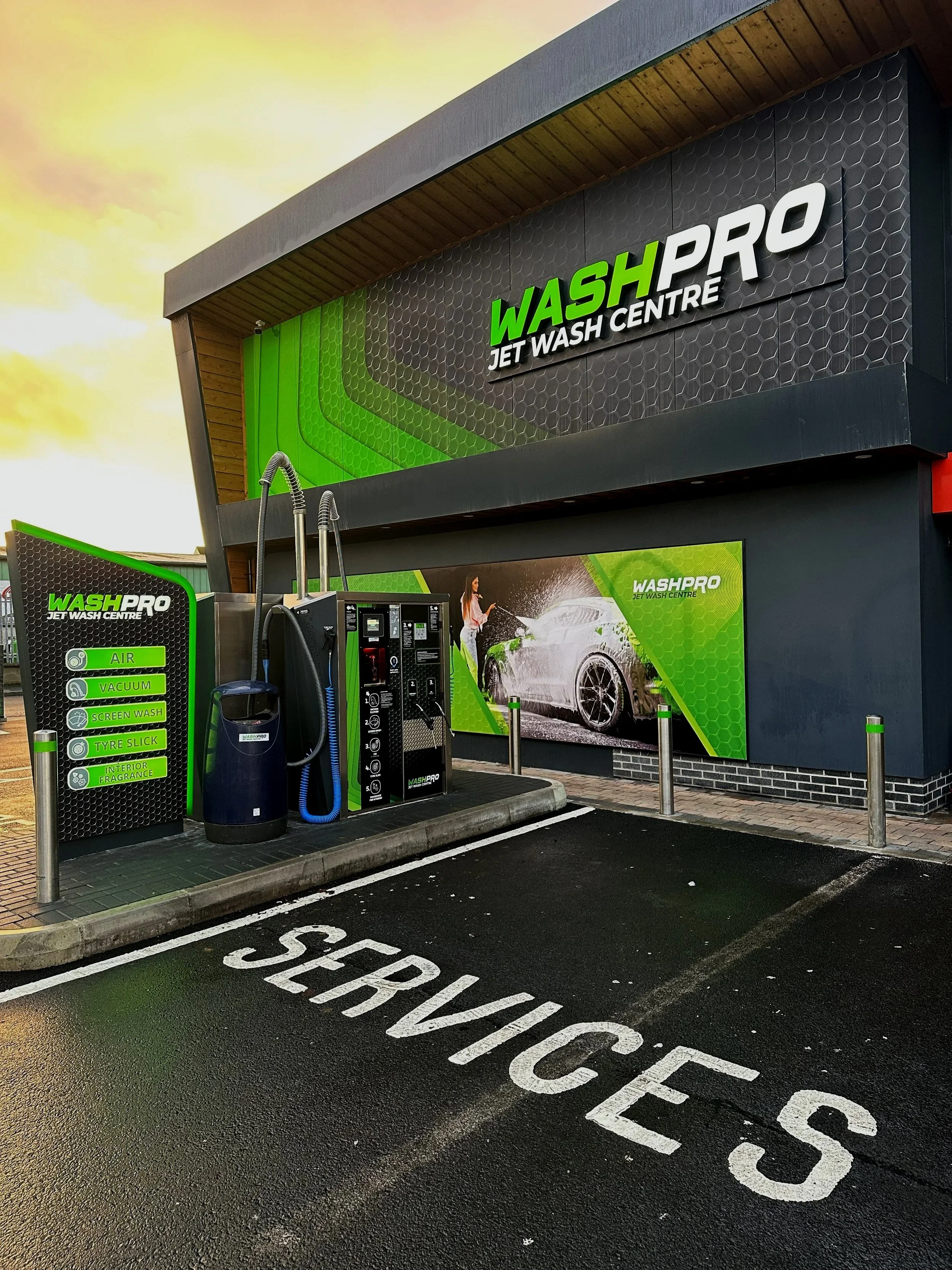 Exterior view of a car wash facility named WASH PRO, with a digital sign, car wash equipment, and a marked parking space labeled 'SERVICE' in white paint on black asphalt.