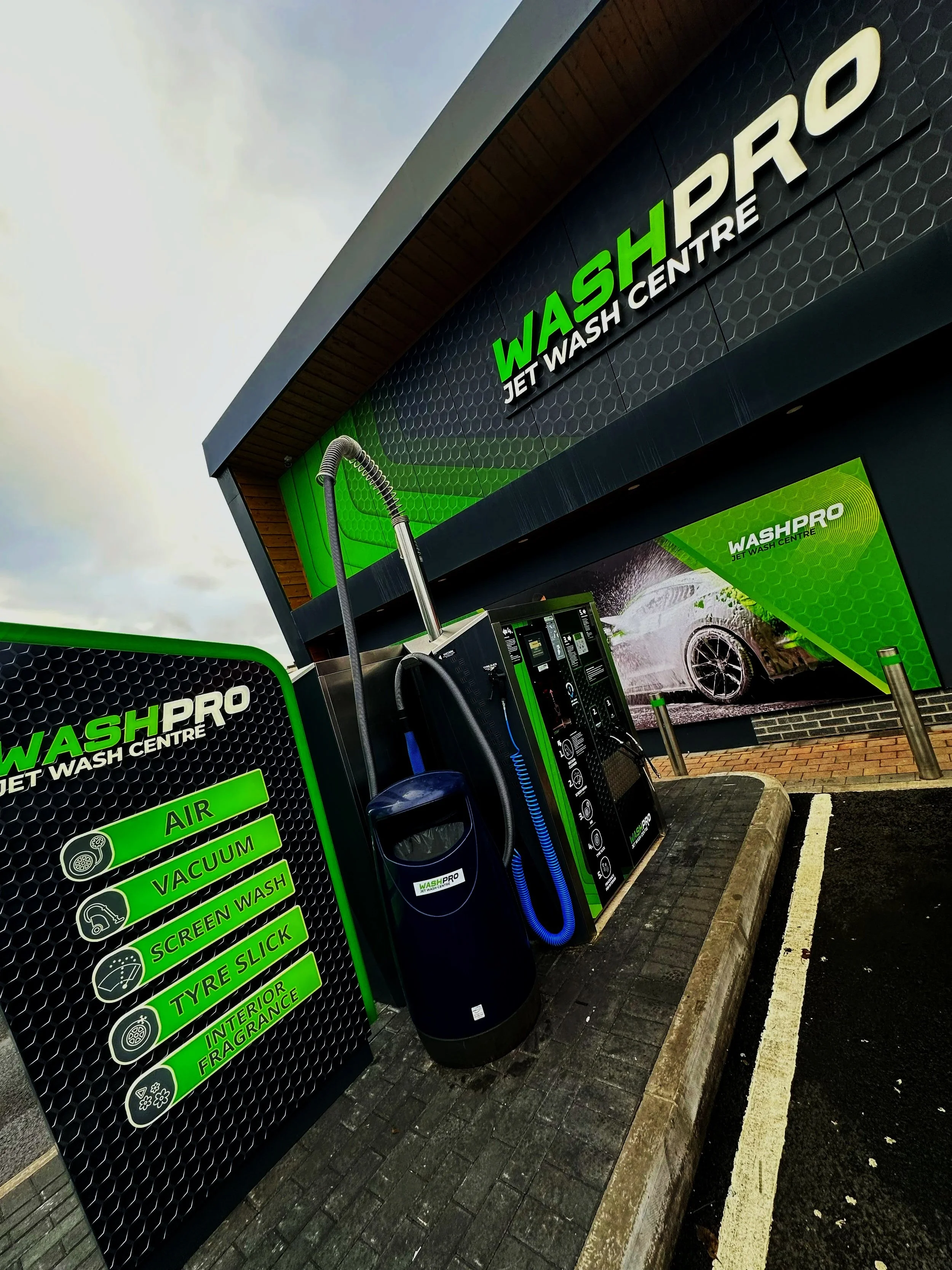 Exterior view of WASHPRO jet wash center showing self-service car wash equipment, a sign listing services such as air, vacuum, screen wash, tyre slick, interior fragrance, and a large photo of a car being washed.