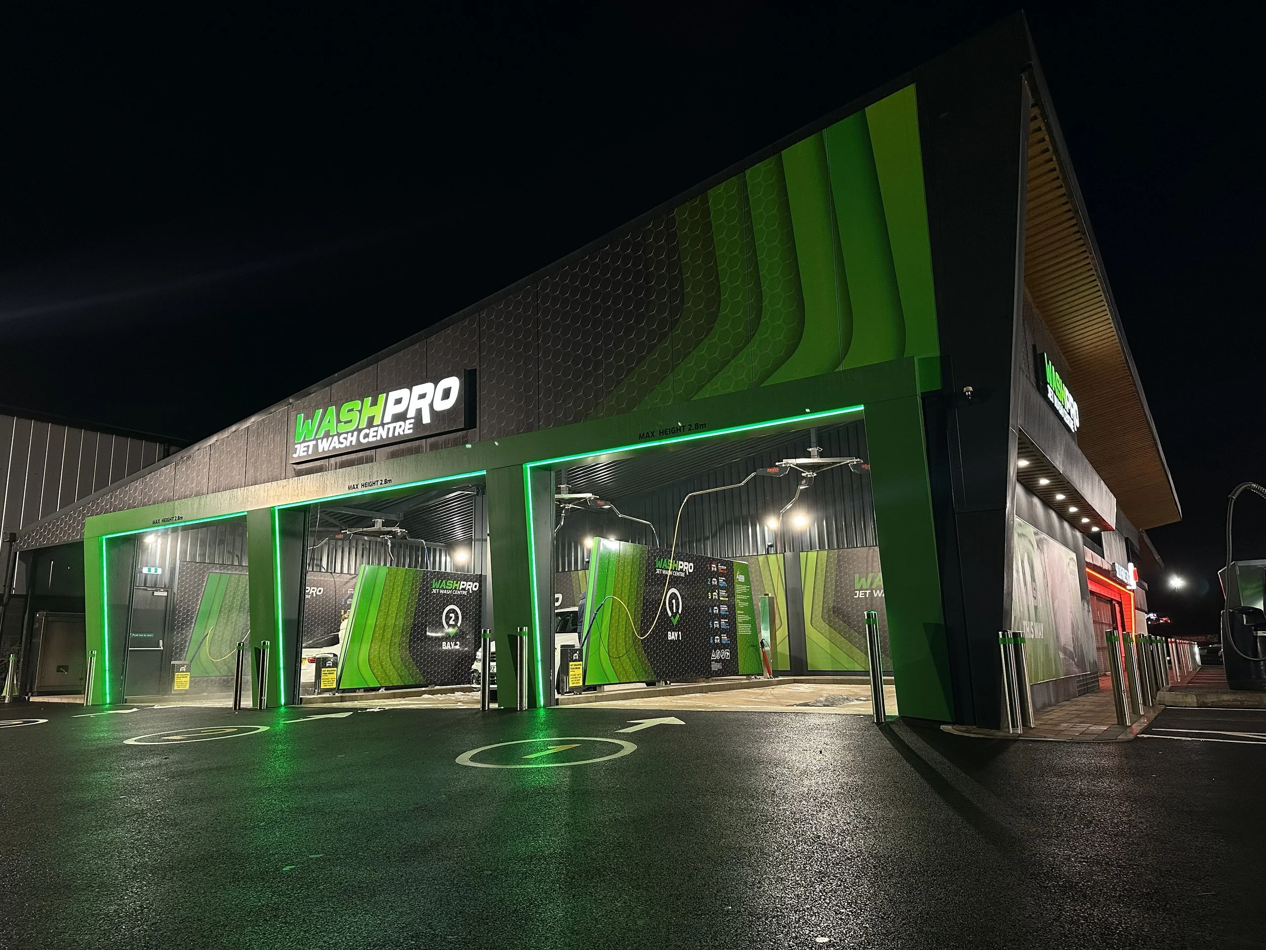 Night view of WashPro Jet Wash Centre with green accents, indoor washing bays, and marked parking spots on wet asphalt.