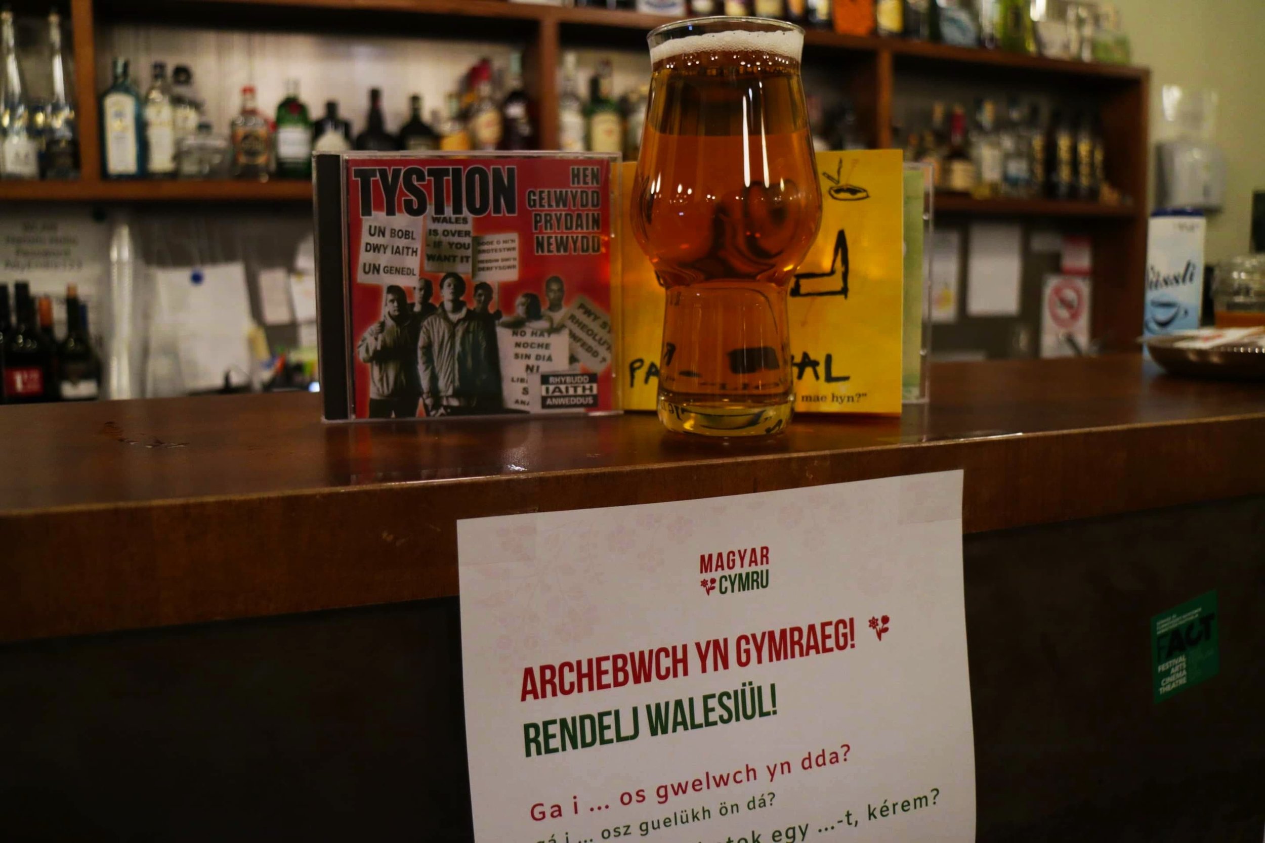 A photo of the bar at Három Holló Café in Budapest, featuring a pint of beer, two Welsh-language CD covers, and a Magyar Cymru-branded sign in Welsh and Hungarian encouraging guests to order in Welsh.