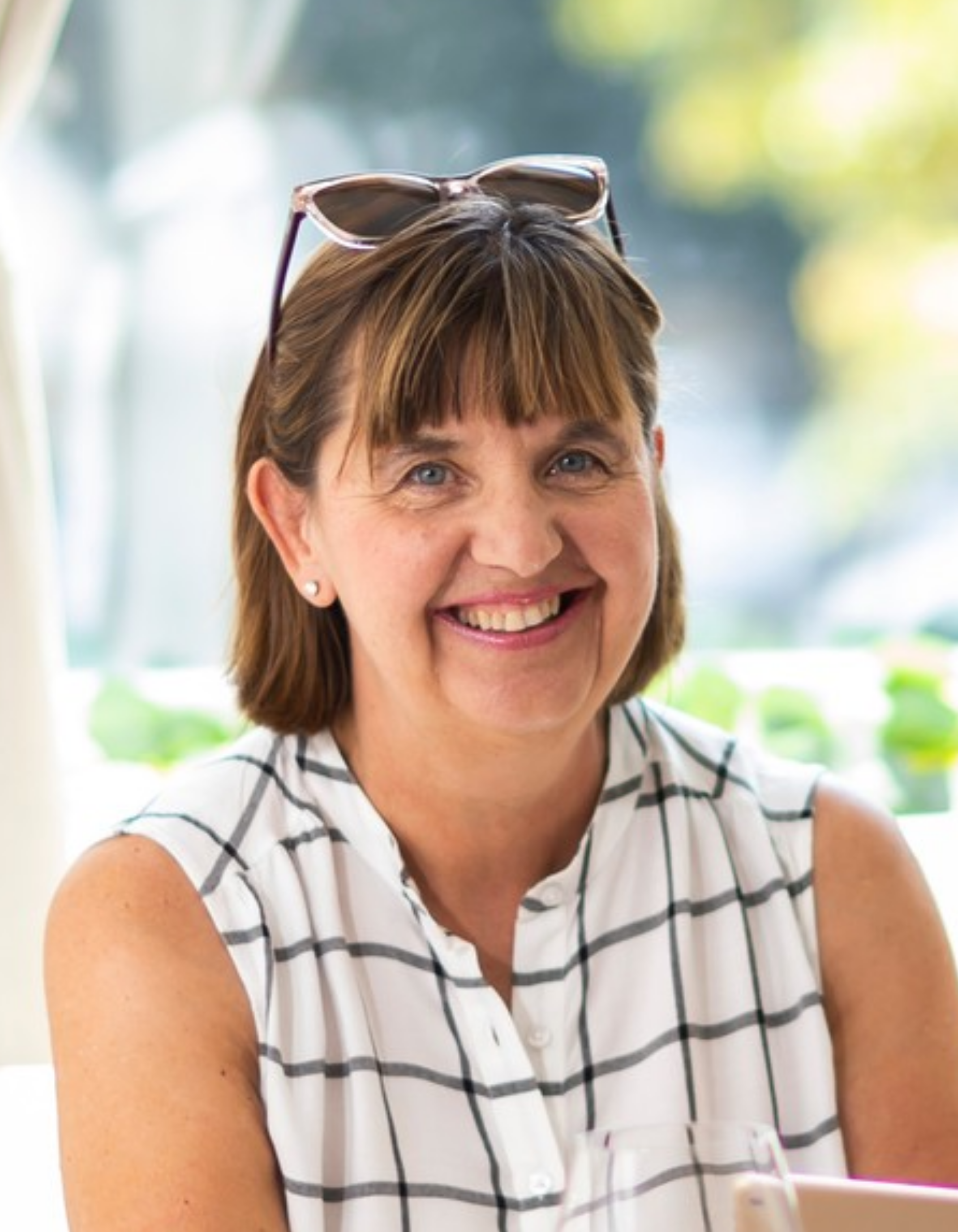 A portrait of director Monika Gyenes wearing a sleeveless white top with black stripes, smiling, with sunglasses resting on her head, sitting indoors near a window with green foliage outside.