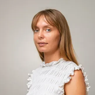 A portait of colleague Gyongyver Jescheta wearing a white top, looking at the camera against a plain gray background.