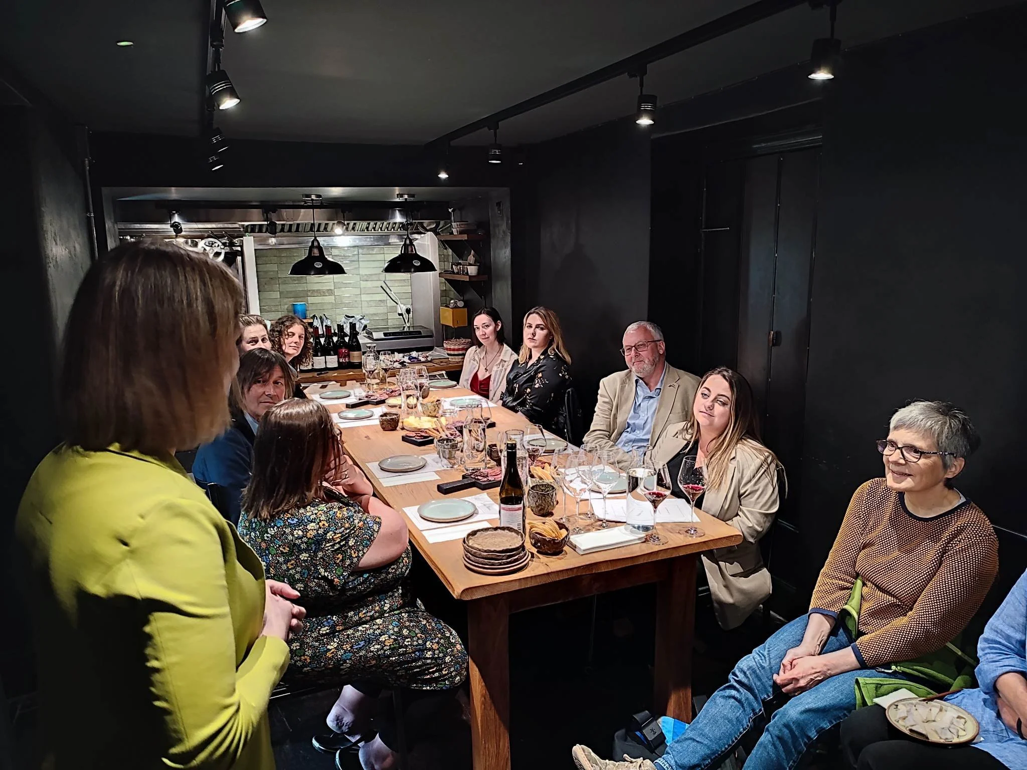 Sommelier Csilla Sebestyén, dressed in green, delivering a Hungarian wine tasting session at Gwen Restaurant in Machynlleth, with several guests sitting around a large table listening to her