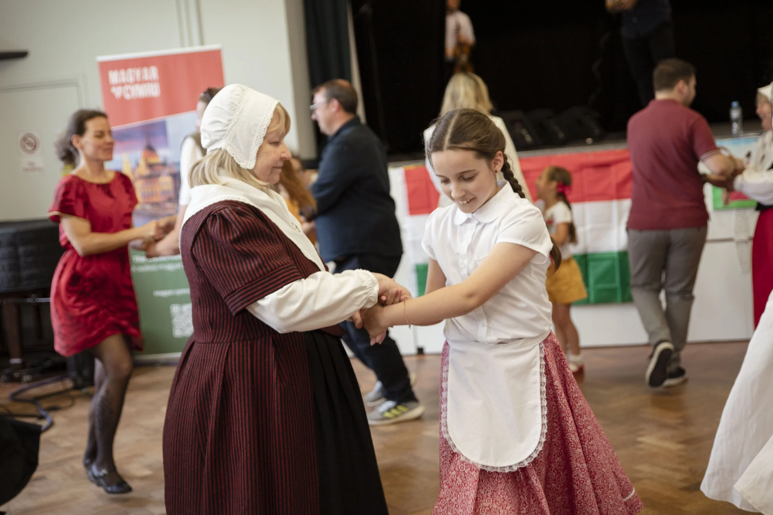 A Welsh folk dancer and a young Hungarian girl in traditional Hungarian folk costume dancing together at an indoor event, other people are dancing and socialising in the background.