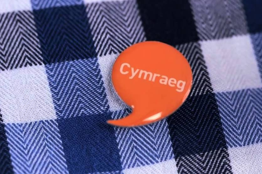 An orange lapel pin in the shape of a speech bubble, featuring the word 'Cymraeg' meaning Welsh, used by Welsh speakers to signal their language skills, placed on blue and white textile