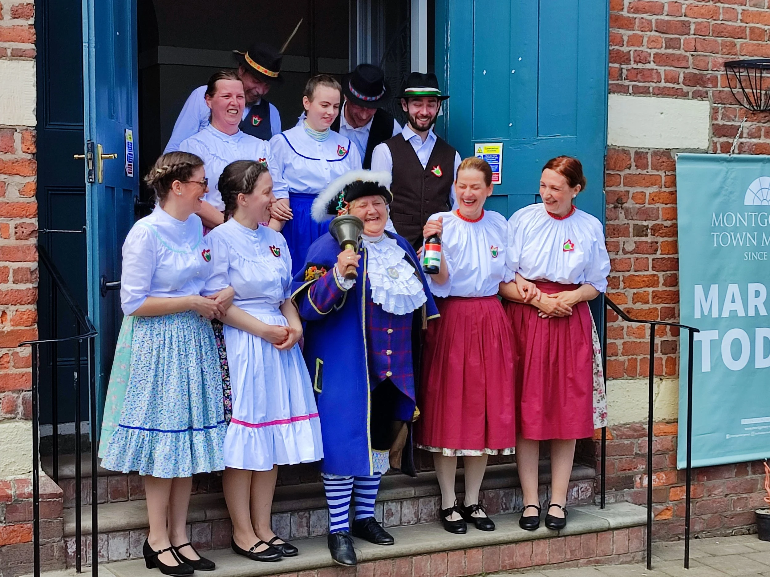 Montgomery town crier Sue Blower, dressed in full regalia while laughing and shaking a bell, posing in front of the town hall with dancers from DunAvon Hungarian Dance Ensemble on Montgomery Hungarian Day.