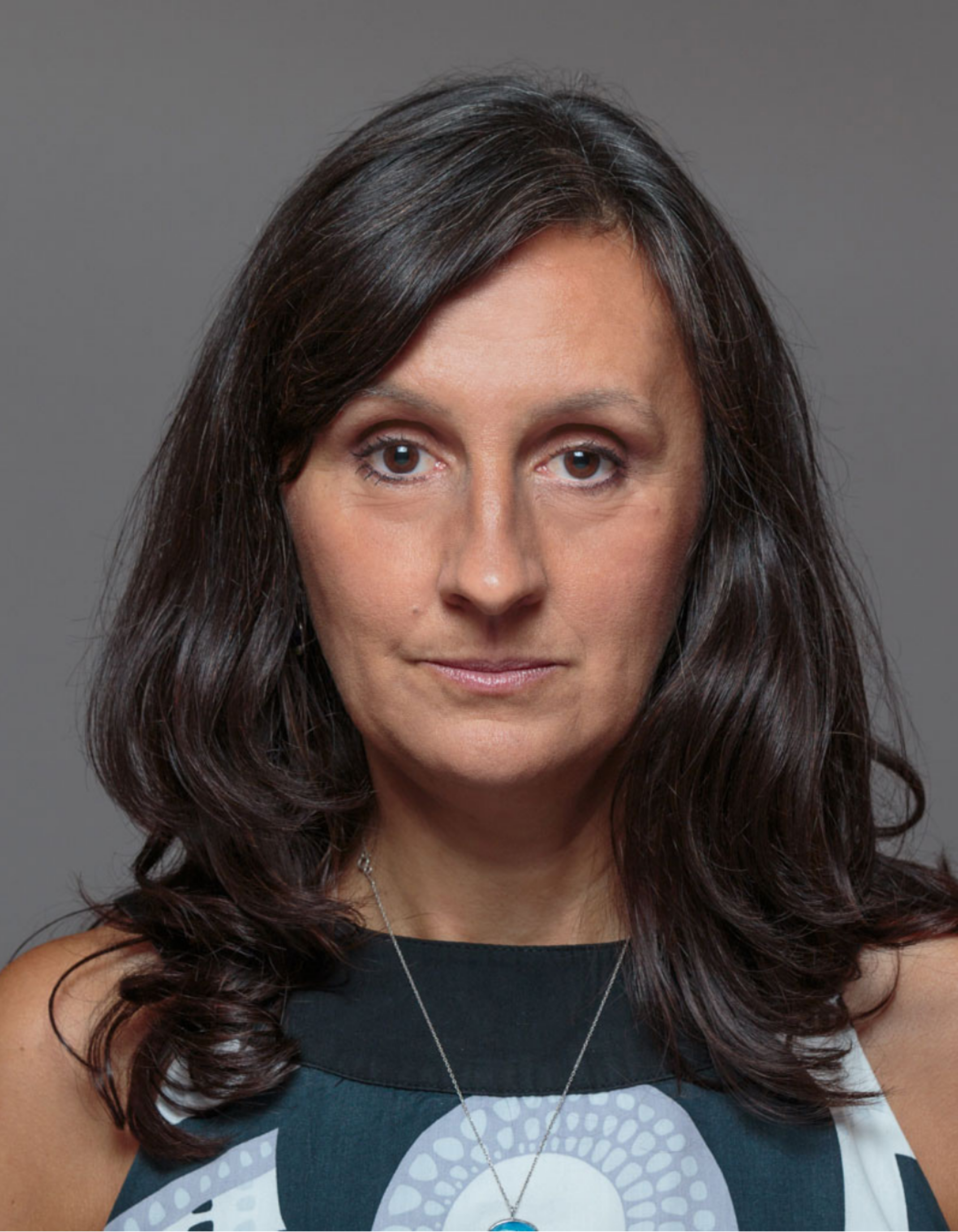 A portrait of director Marta Kecskes wearing a sleeveless top with a black and white pattern and a silver necklace with a blue pendant.