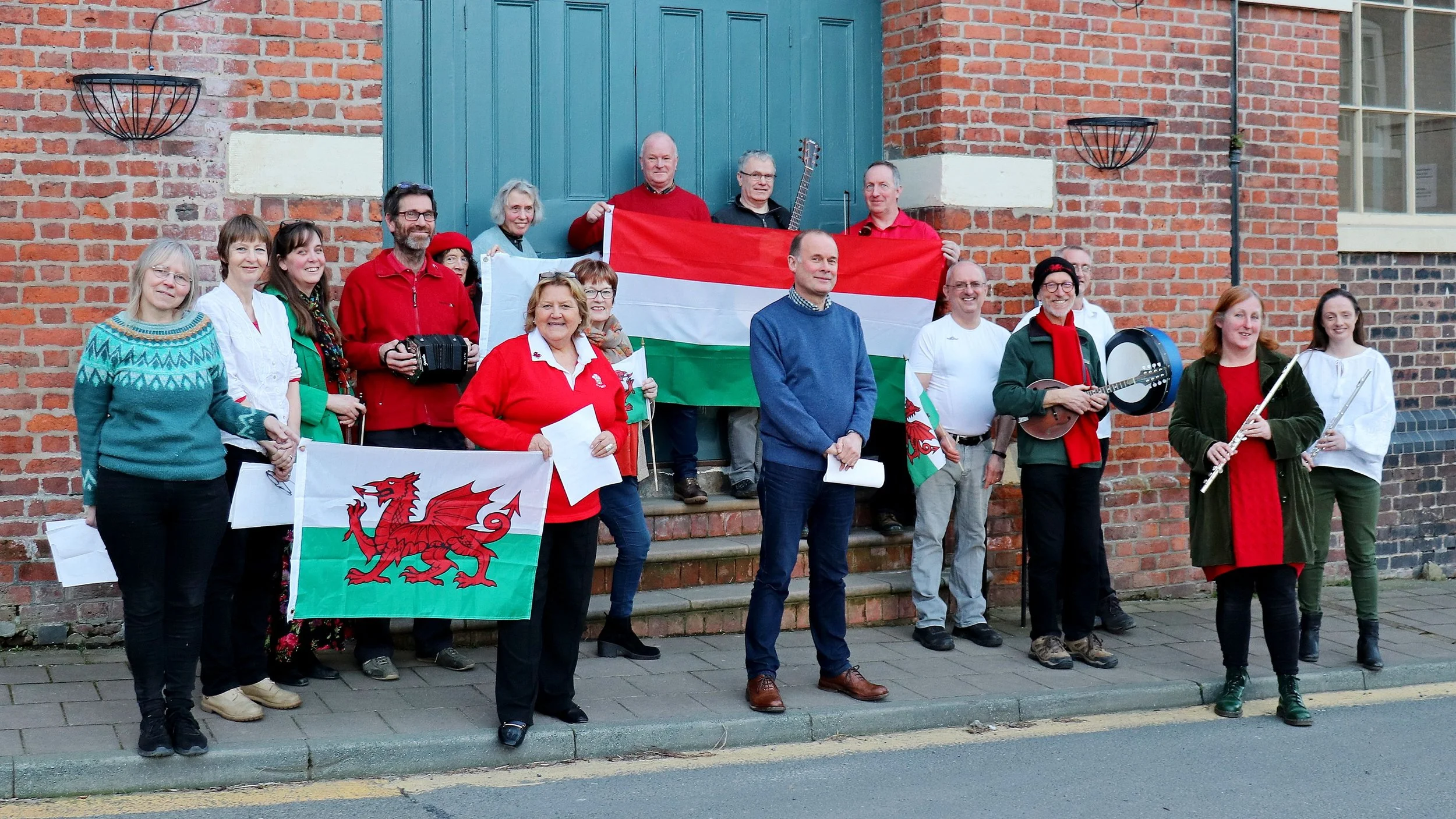Residents of Montgomery in Wales dressed in red, white, and green, standing in front of the town hall, holding Welsh and Hungarian flags and musical instruments, celebrating cultural friendship between their Welsh town and Hungary.
