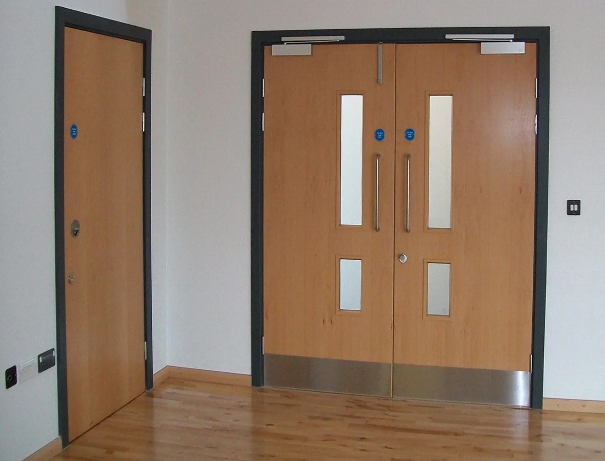 Wooden double doors with windows and metal rail at the bottom, and a single wooden door with windows to the left, all with black frames, in a room with white walls and wooden floors.