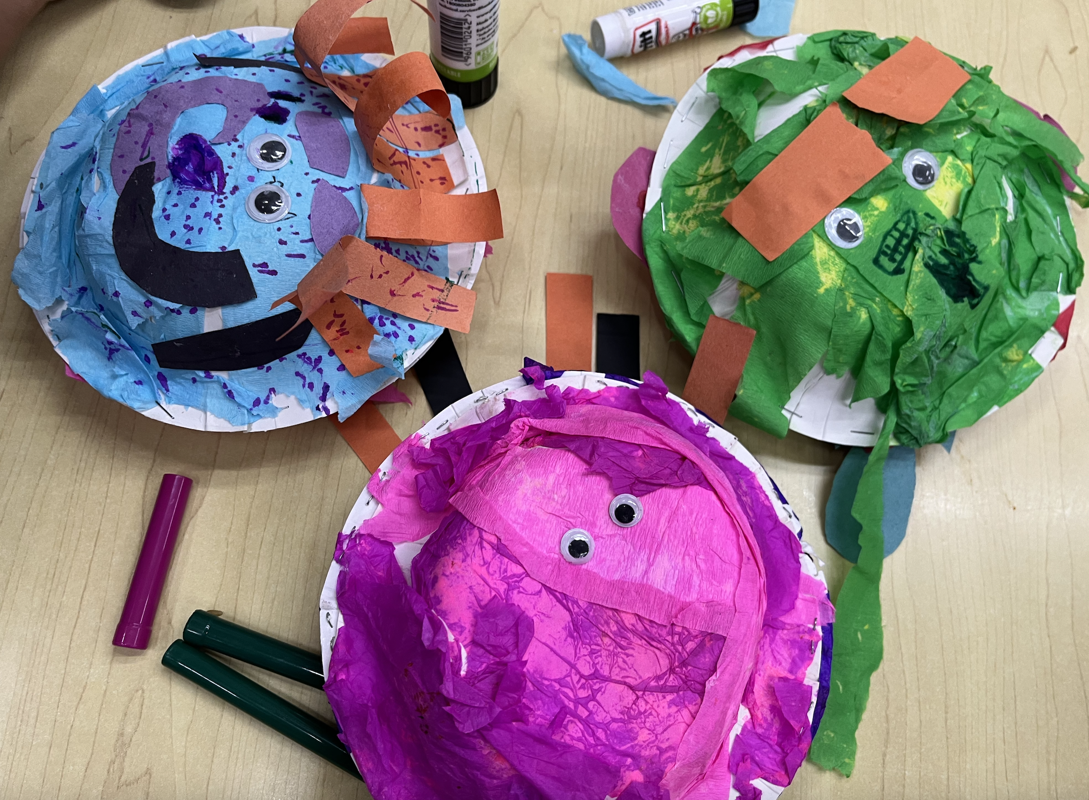 Three colorful decorated paper plate masks with googly eyes, placed on a wooden table surrounded by art supplies including markers, glue, and tissue paper.