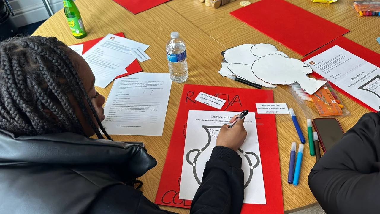 Person with braided hair writing on a worksheet at a table with red folders, pens, papers, a water bottle, and some cutouts.