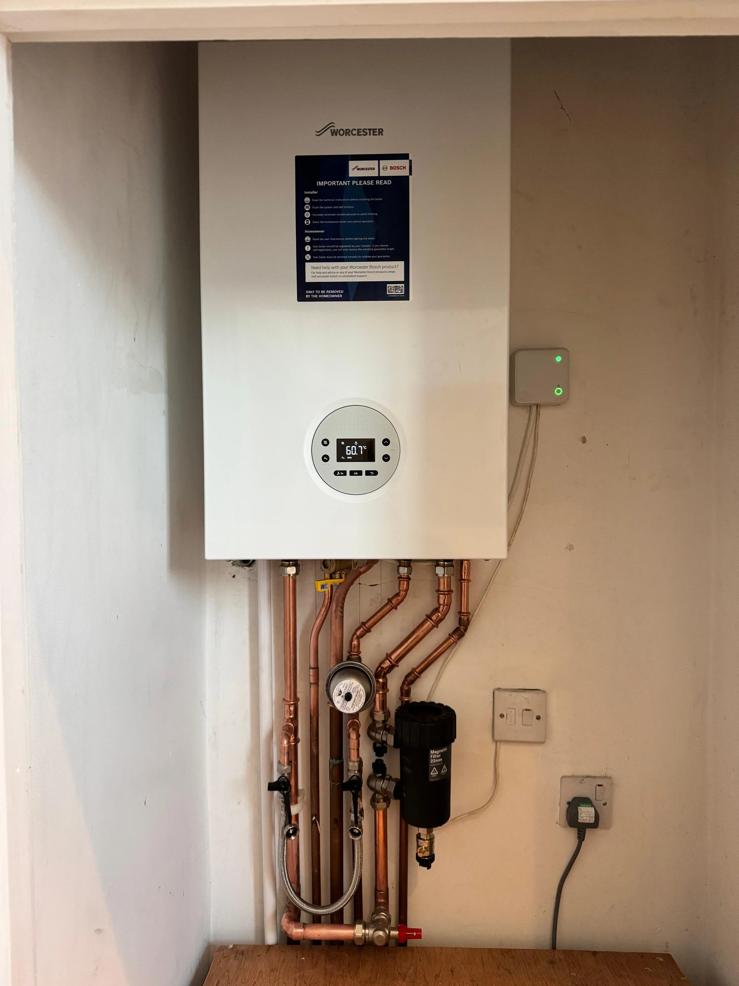 Wall-mounted white boiler system with digital display showing 60.7°F, connected to copper pipes, with a nearby electronic control box plugged into an outlet.