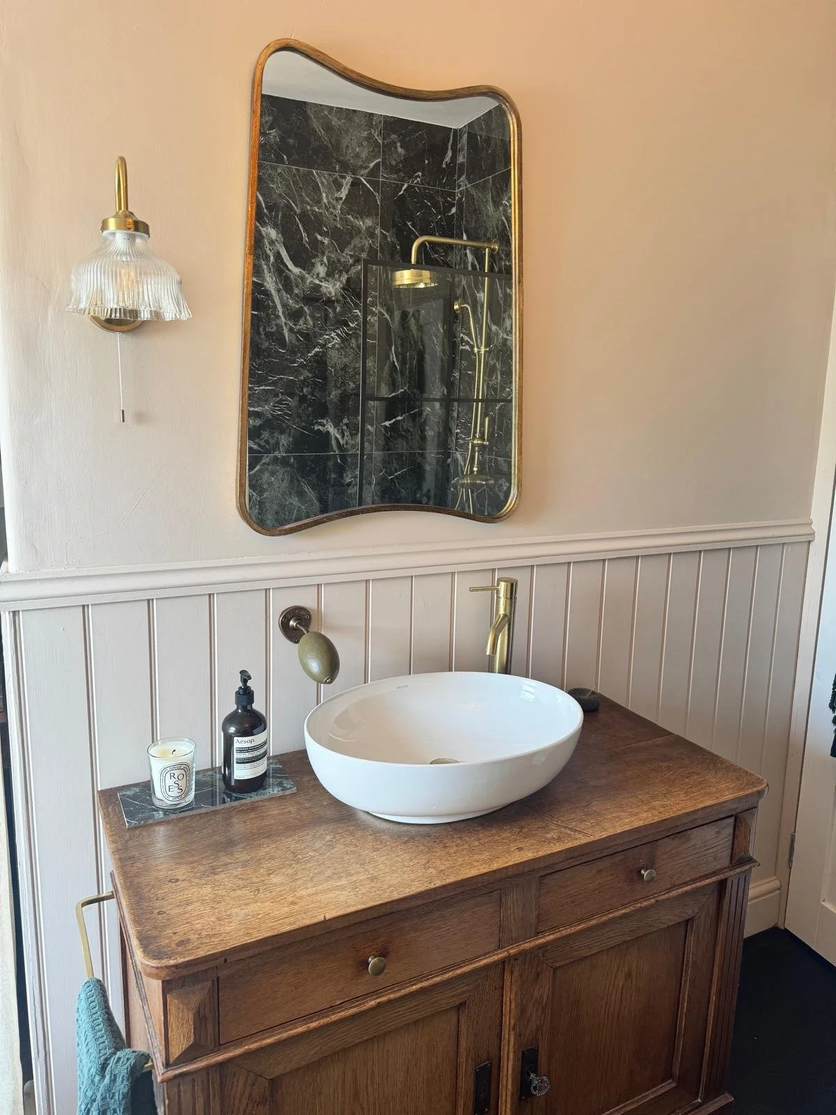 Bathroom vanity with a round white vessel sink, brass faucet, a wooden cabinet, and a mirror with wavy edges. The bathroom has beige walls with wainscoting, a wall-mounted light fixture, and a soap dispenser on the countertop.