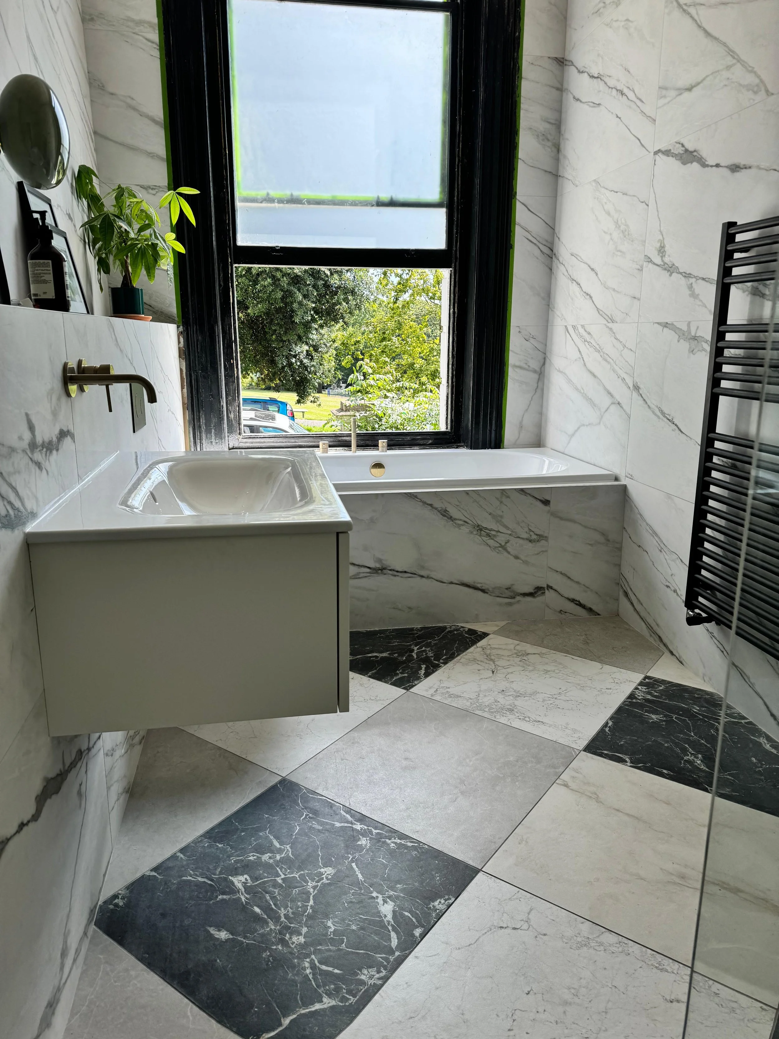 Modern bathroom with marble tiles, a large window overlooking greenery, a white bathtub, floating vanity with sink, black towel radiator, and a potted plant.