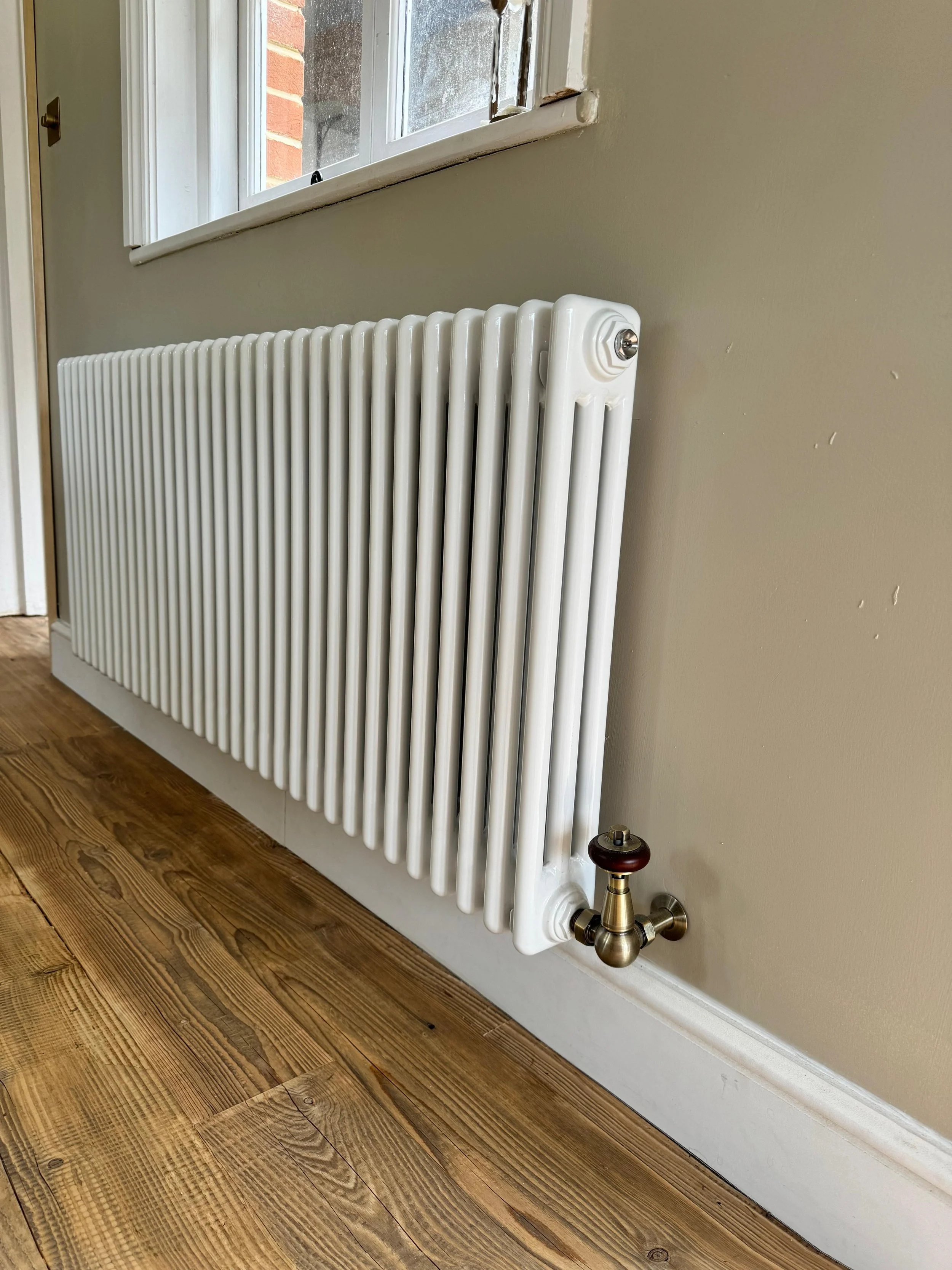 White radiator mounted on a beige wall near a wooden floor, with a window above.