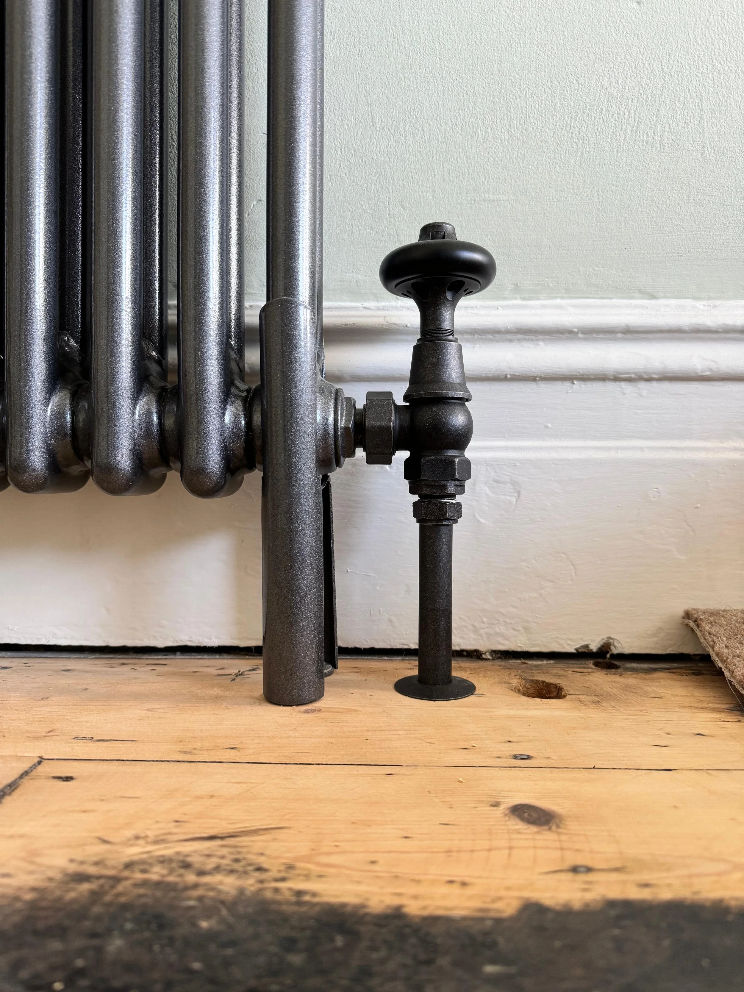 Close-up of an old black radiator pipe connected to a radiator, with a wooden floor and white wall in the background.