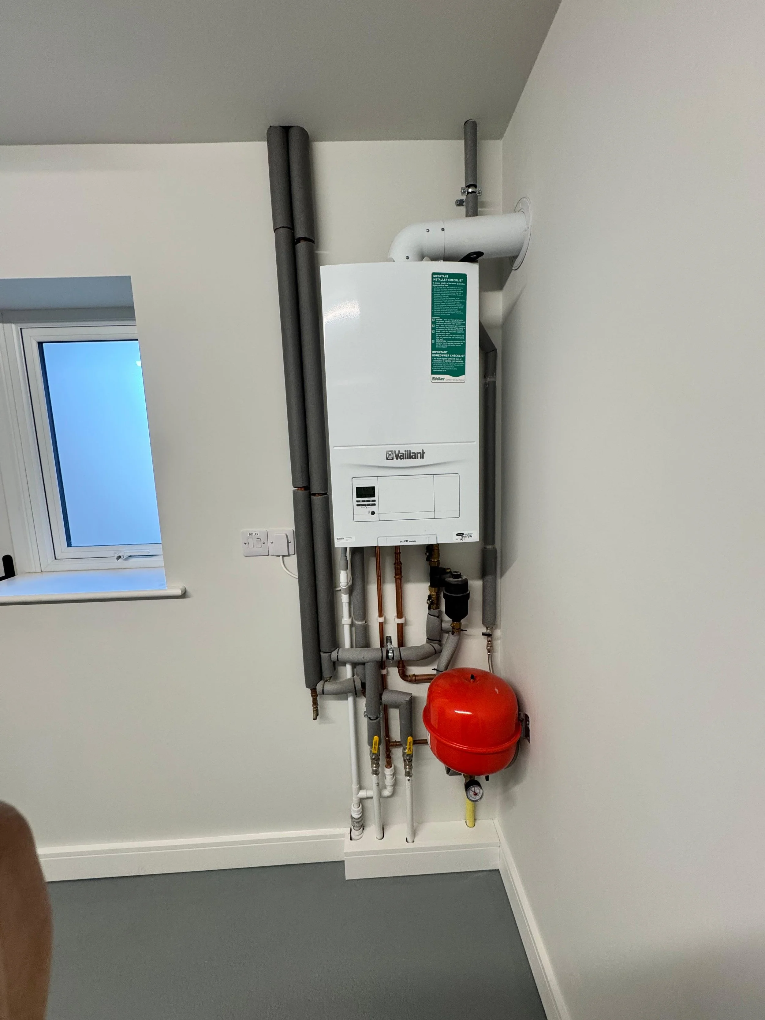 A white wall-mounted boiler system with pipes and a red expansion tank in a room with a window, gray flooring, and white walls.