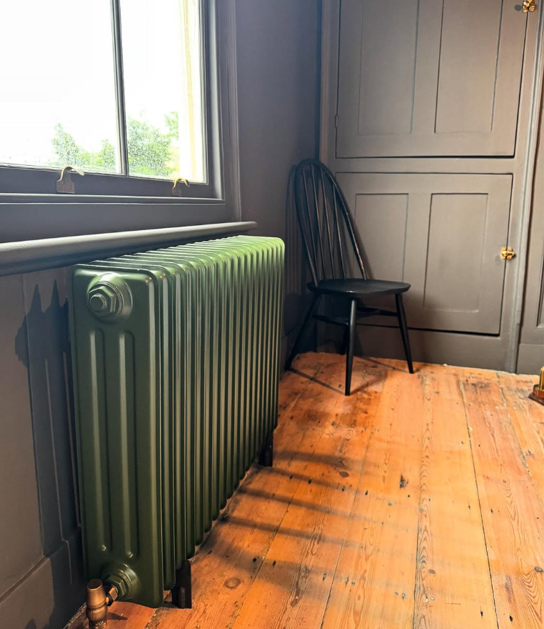A room with a green radiator, a black chair, and a gray closet next to a window with a view of greenery outside. The room has wooden flooring and soft natural light.