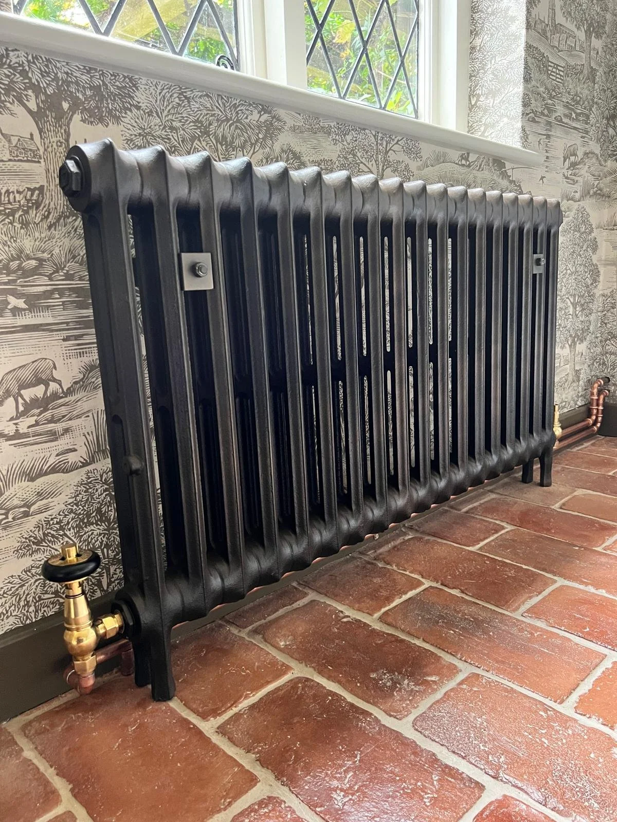 A black cast iron radiator against a wall with wallpaper depicting nature scenes, positioned on a red brick floor near a window with a leaded glass pattern.