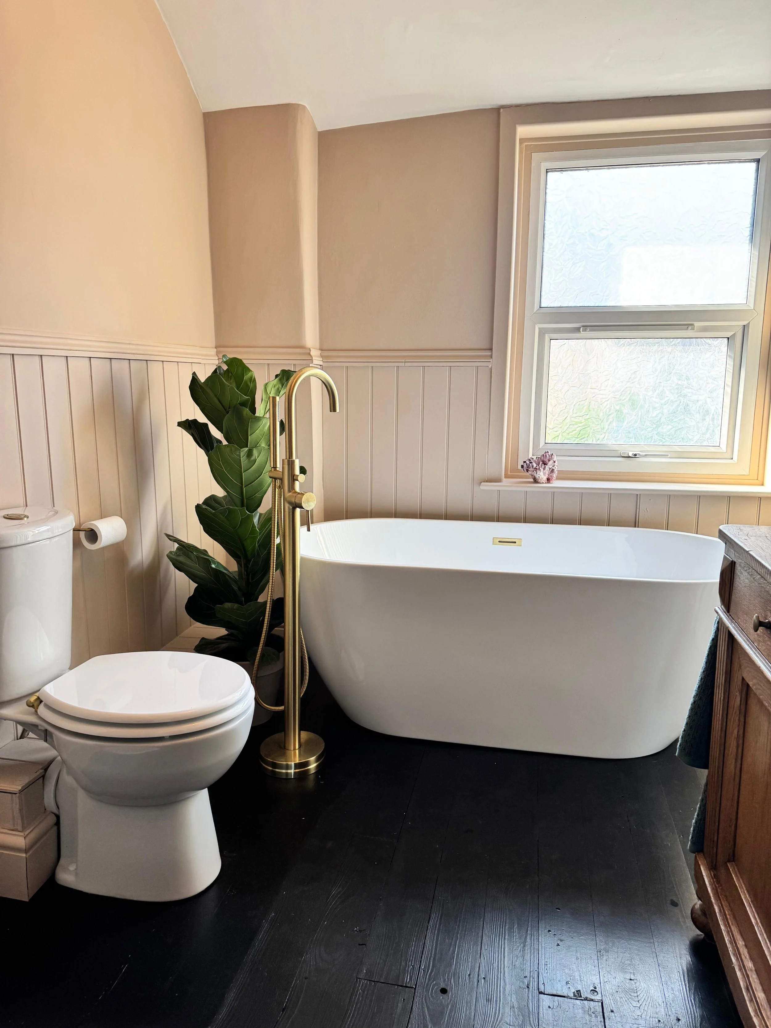 A bathroom featuring a white modern toilet, a black floor, a large white bathtub beside a window, a green plant next to the bathtub, and a wooden cabinet on the right.