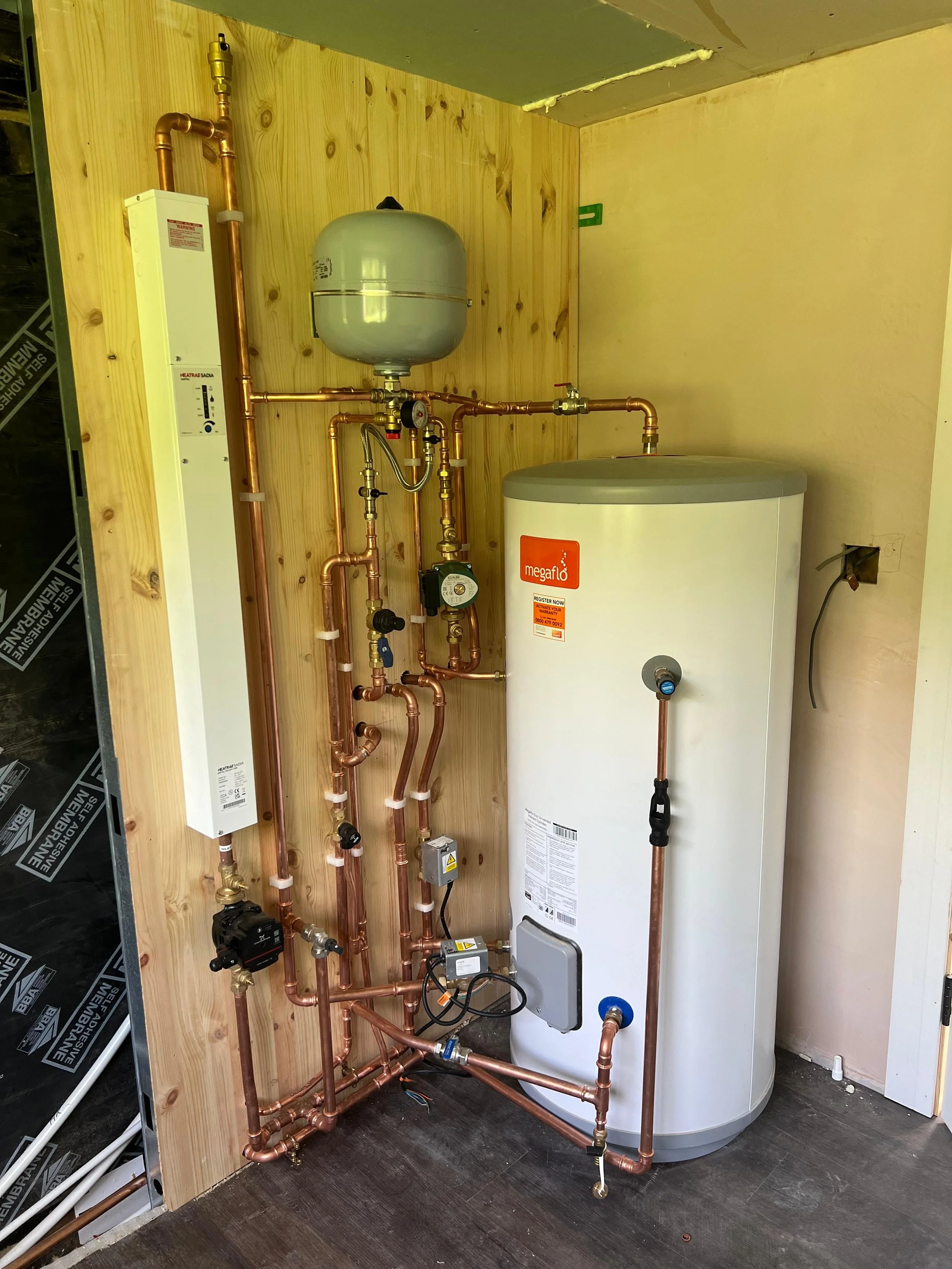 A water heater with copper piping, pressure regulators, and a expansion tank installed on a wooden wall in a utility room.