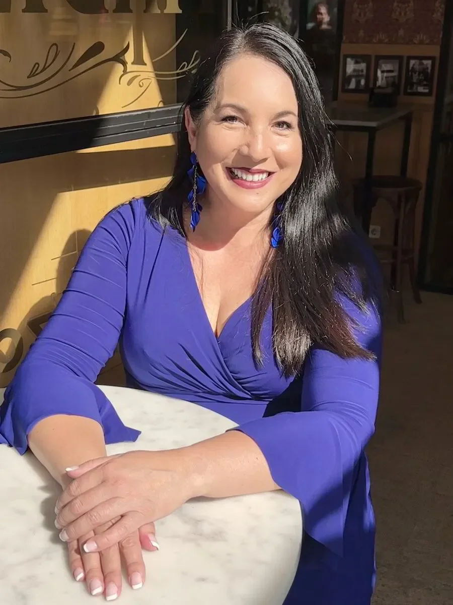 A woman with long black hair, wearing a royal blue dress and matching earrings, sitting at a table with her hands folded, smiling in a warmly lit indoor setting.