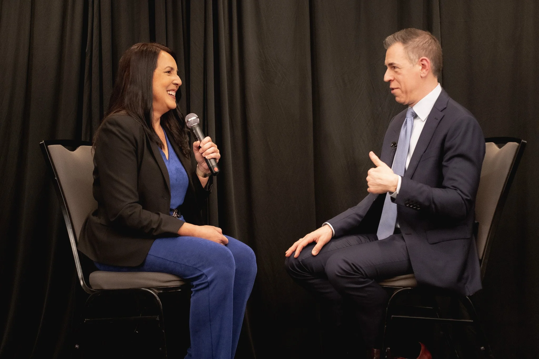 Two people sitting on chairs facing each other, engaged in conversation on a stage with black curtains in the background. The woman on the left is holding a microphone and smiling, wearing a black blazer and blue outfit. The man on the right is wearing a dark suit and light tie, listening attentively with a slight smile.