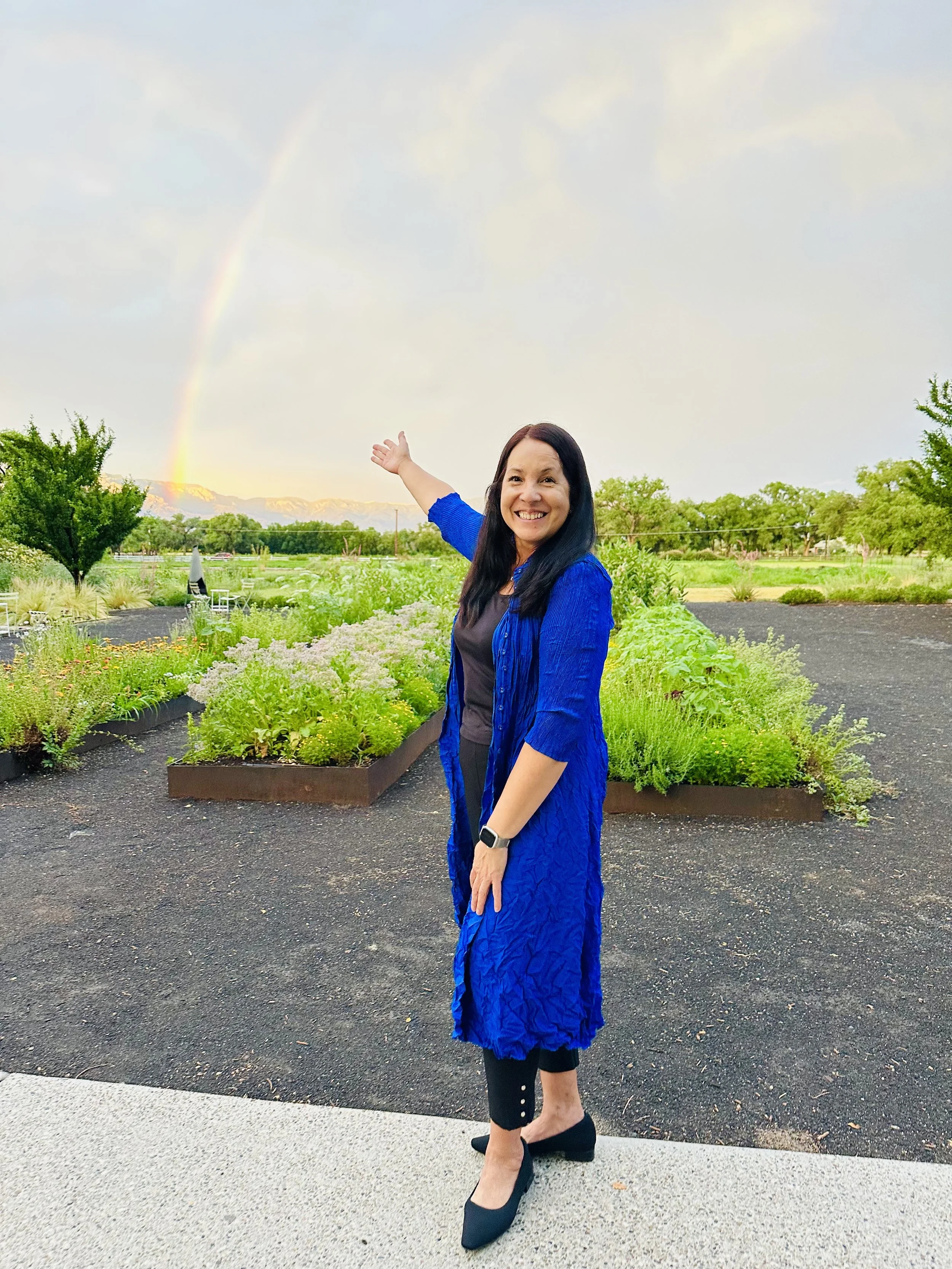 Woman in a blue coat smiling and pointing at a rainbow in the sky behind a garden and landscape.