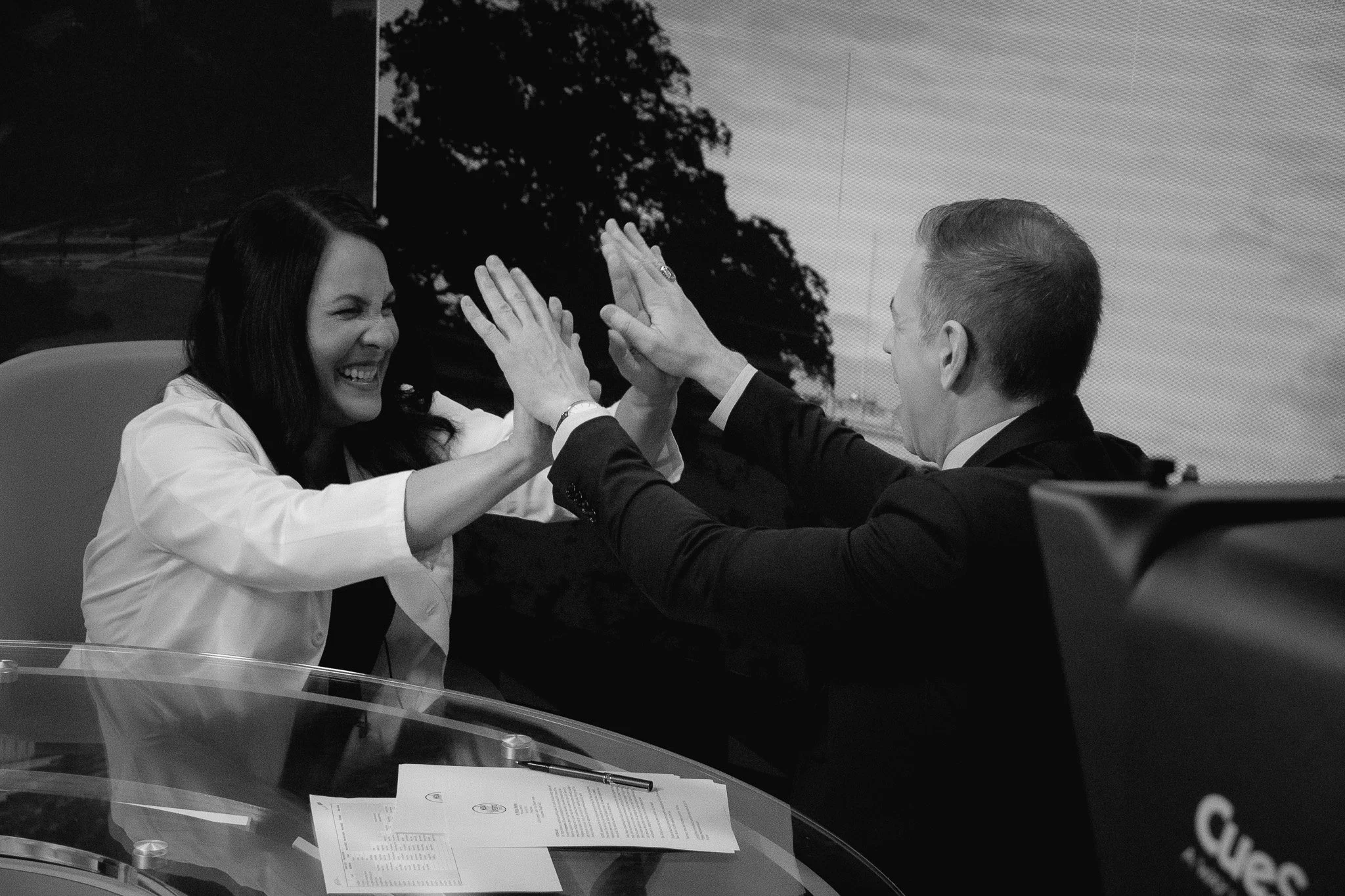 A woman and a man, both smiling and laughing, give each other a high five across a table with documents and a pen on it, in a professional setting.