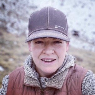 Person wearing a baseball cap and a brown jacket outdoors with snow-covered trees in the background.