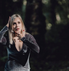 A young woman with long blonde hair in a forested area, making a surprised or confused facial expression while holding her head with one hand.