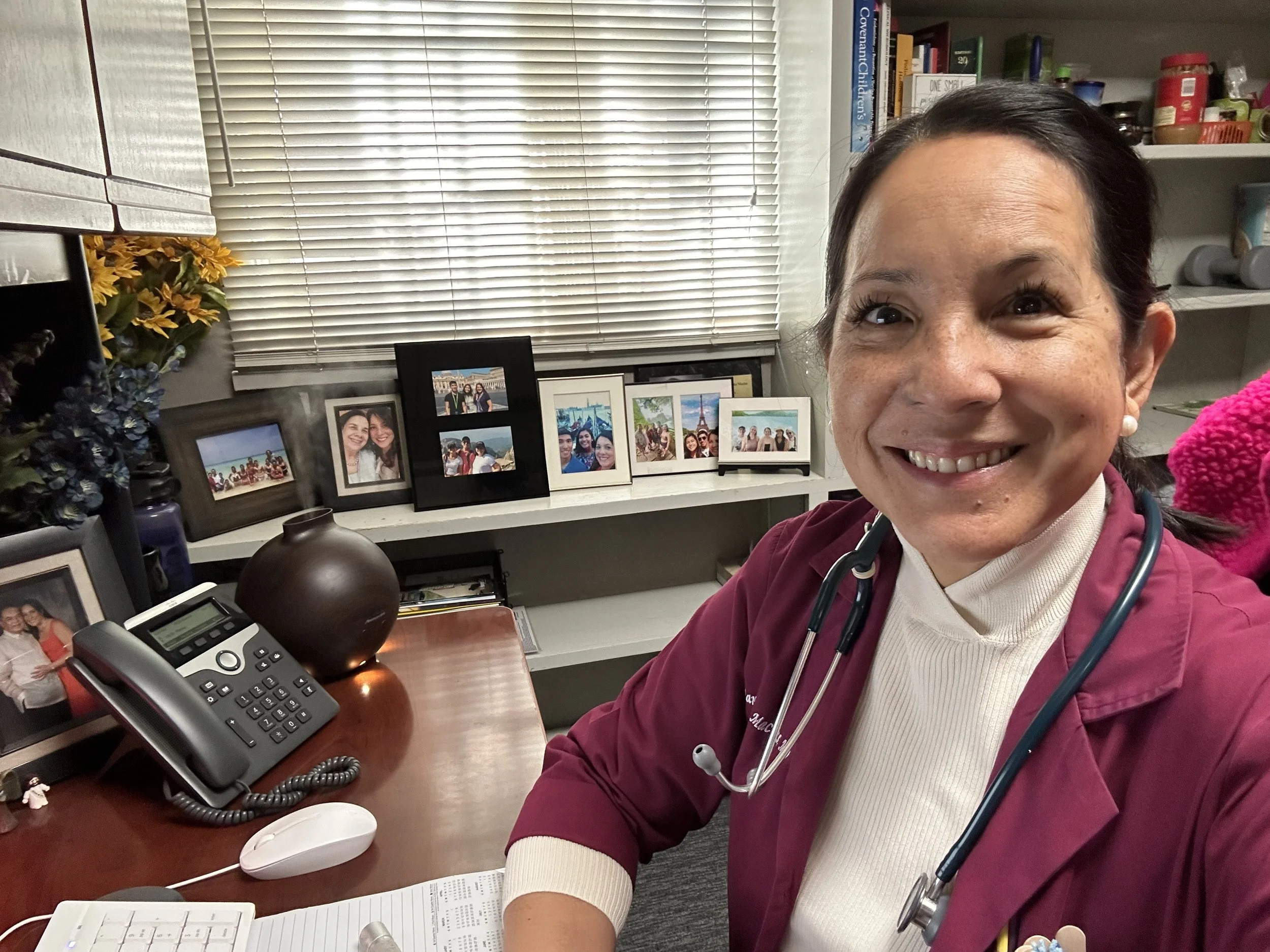 A smiling female healthcare professional wearing a maroon scrub top with a stethoscope around her neck taking a selfie in her office. Behind her are framed photos on a white shelf, a window with blinds, and various books and supplies on shelves.