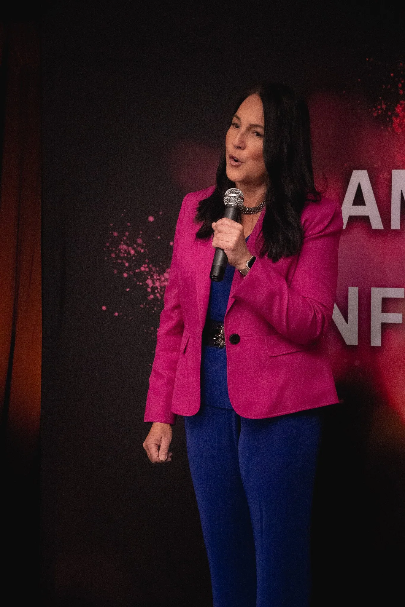 Woman in pink blazer speaking into a microphone at an event with a black background and pink splashes.