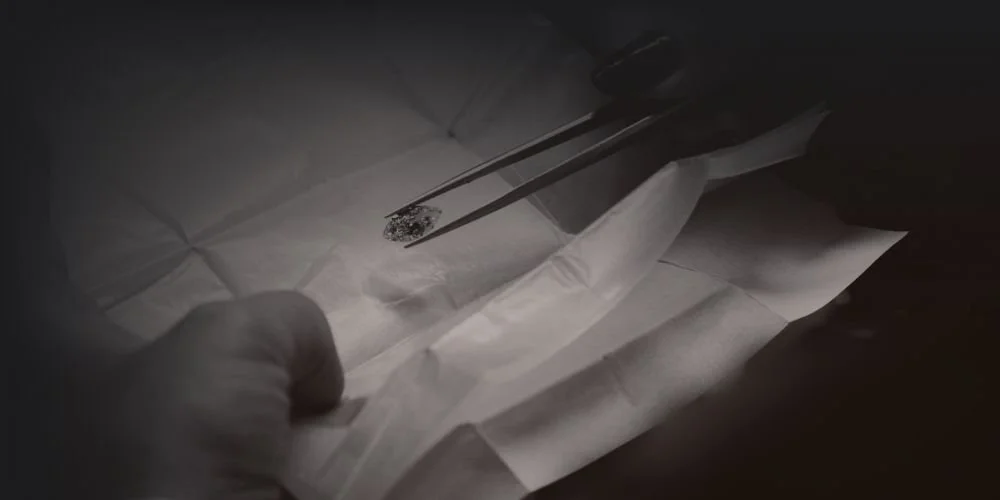 Close-up of a hand wearing a glove using tweezers to handle a small amount of a dark, granular substance on a piece of white paper in a dark environment.