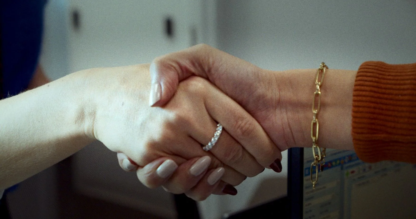 Close-up of two hands shaking, one hand with a diamond ring and a gold bracelet, and the other with painted nails, against a blurred background.