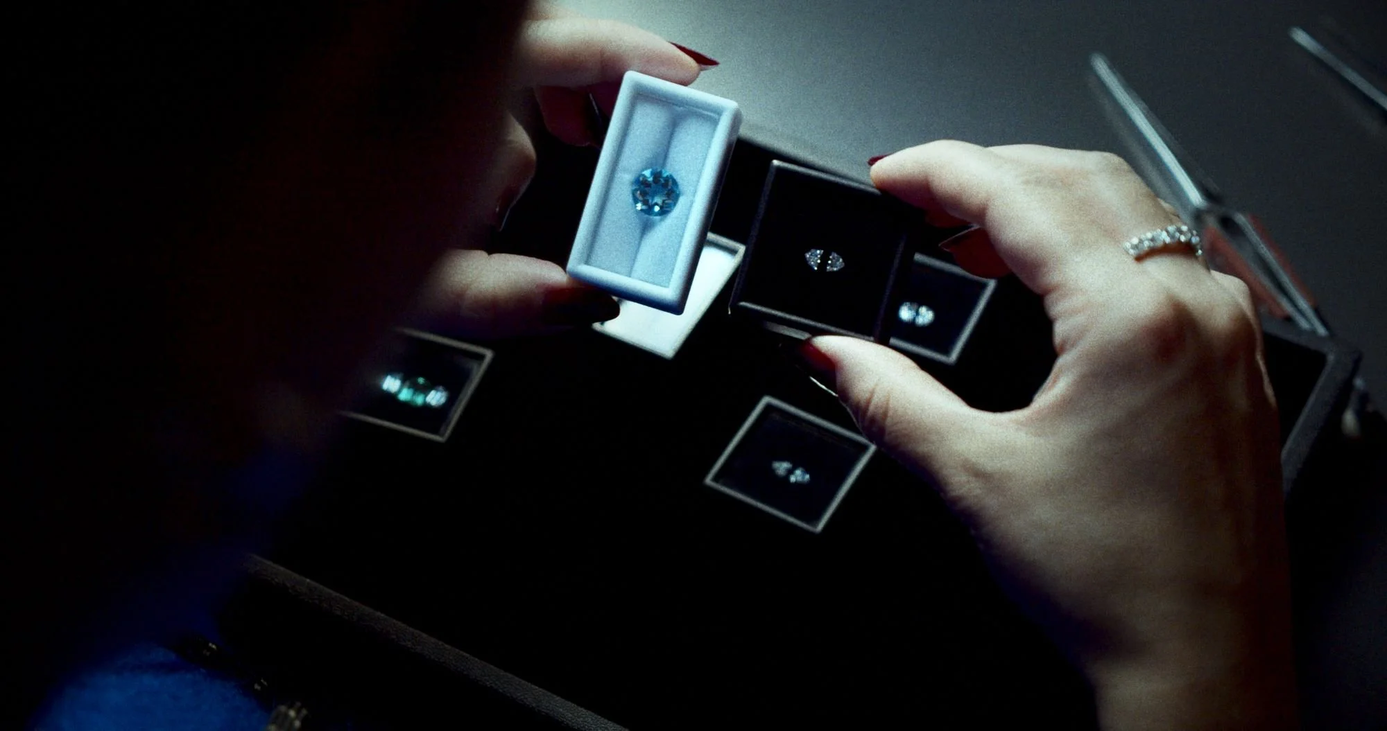 A person holding a light blue gemstone ring in a jewelry box, surrounded by other jewelry boxes containing rings, in a display case.