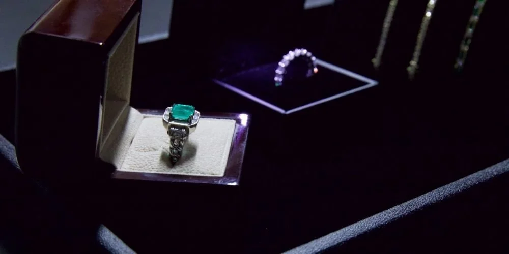 Close-up of a jewelry box with a silver ring featuring a large green gemstone, near other jewelry items like a bracelet and a pair of earrings, displayed in a dark setting.