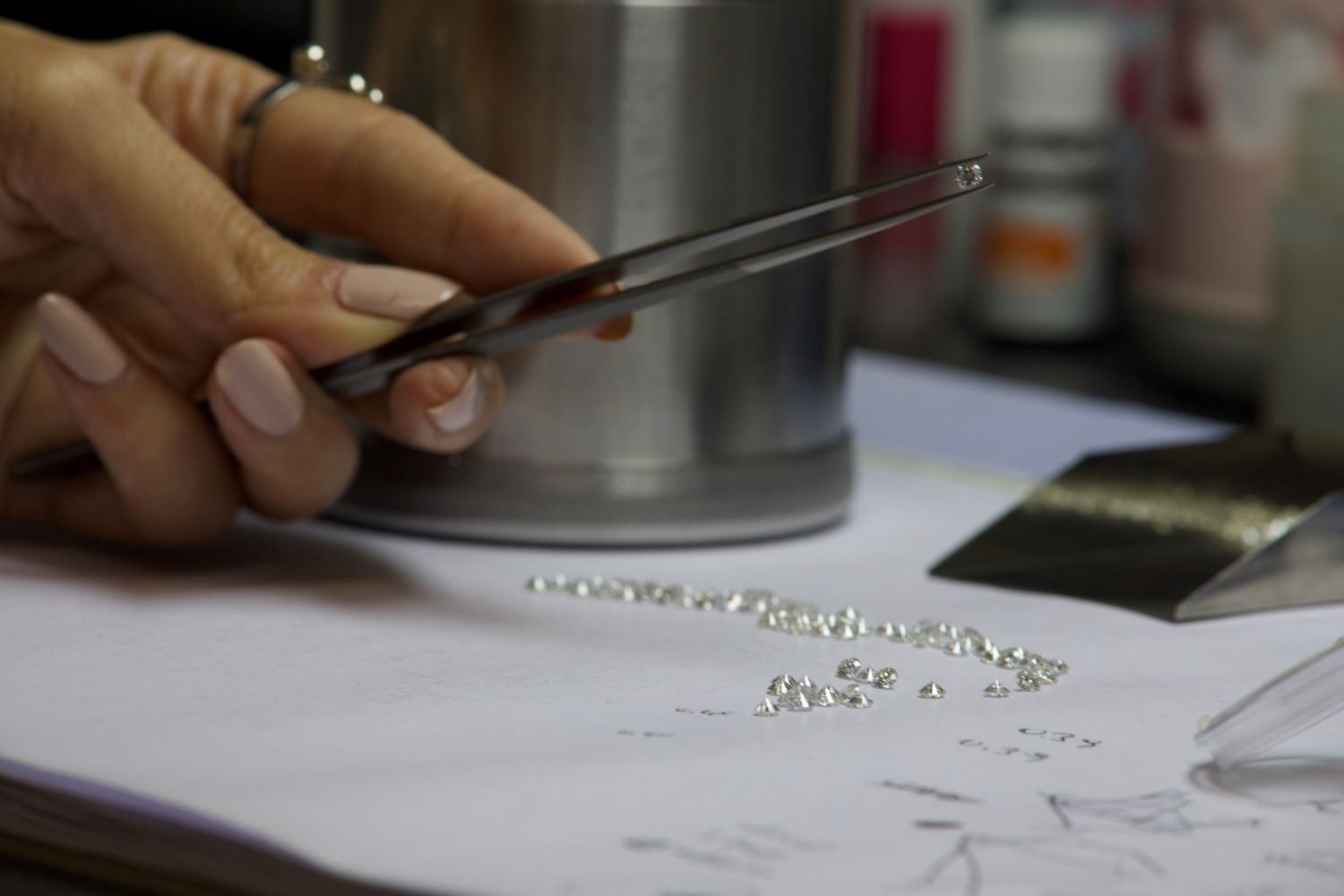 Close-up of a person’s hand holding tweezers, working with small, shiny gemstones or beads on a white work surface.