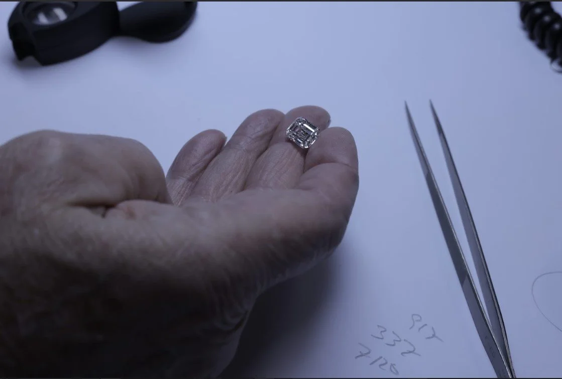 Close-up of a hand with a large, ornate diamond ring on the finger, next to a pair of tweezers and a handwritten note on a white surface.