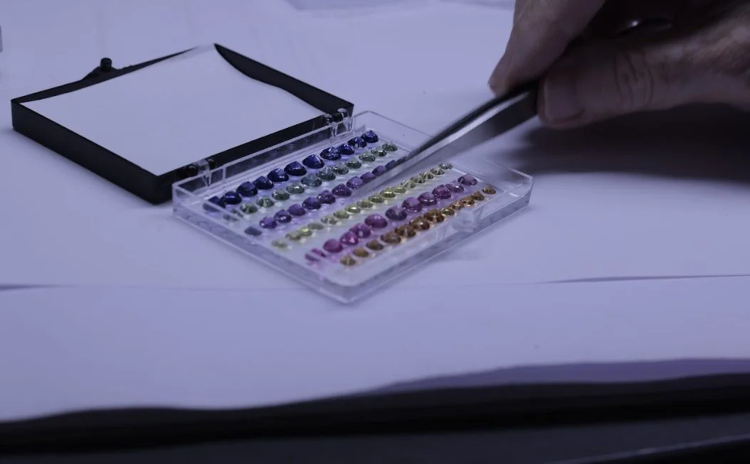Close-up of a hand using tweezers to handle small, colored gemstones or cabochons arranged in rows inside a plastic storage case with a black lid, placed on a white surface.
