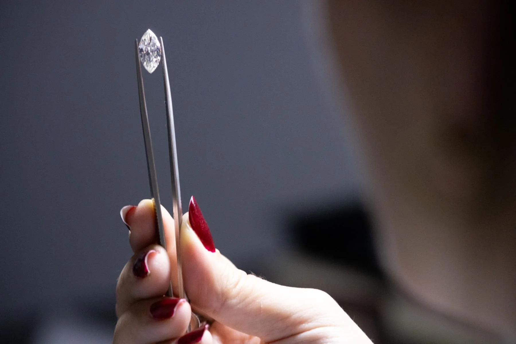 A hand with red painted nails holding a pair of tweezers with a diamond gemstone at the tip.